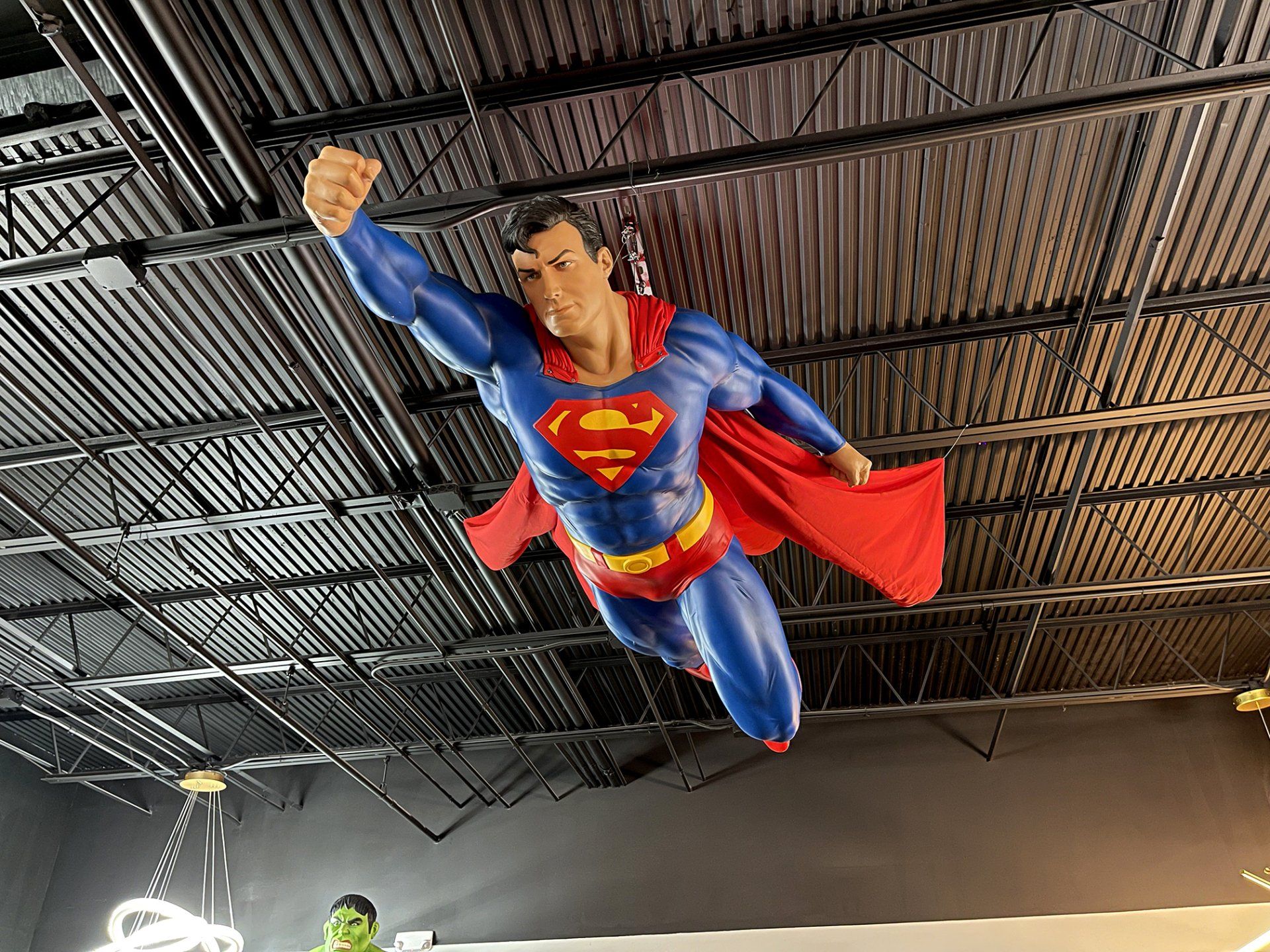 Superman figure flying indoors under a metal ceiling, wearing blue suit and red cape