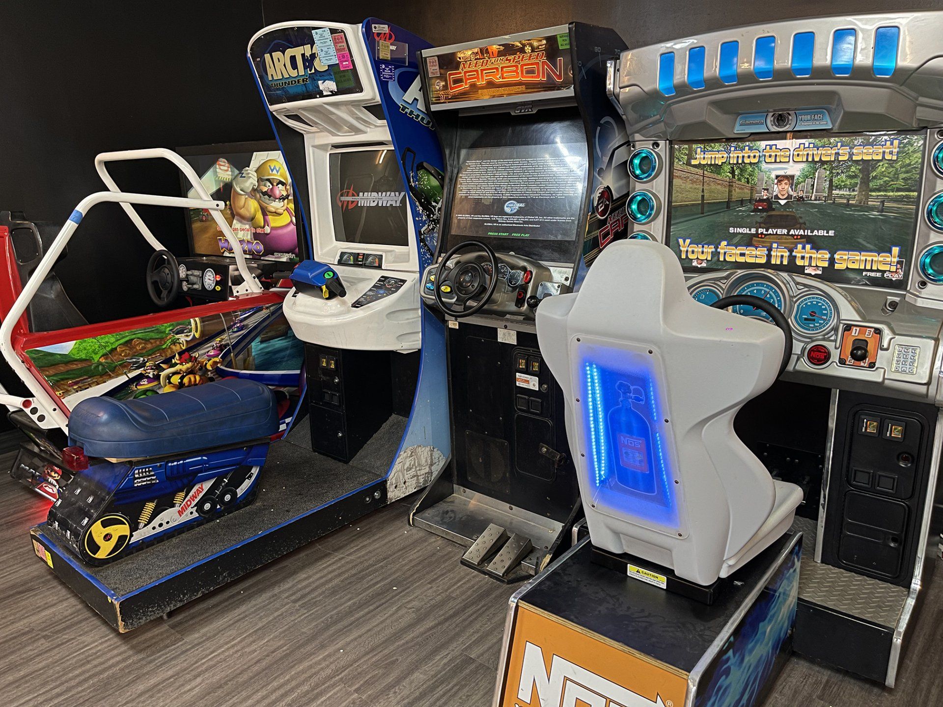 Arcade racing game cabinets with blue-lit seats and a driving simulator screen in a game room