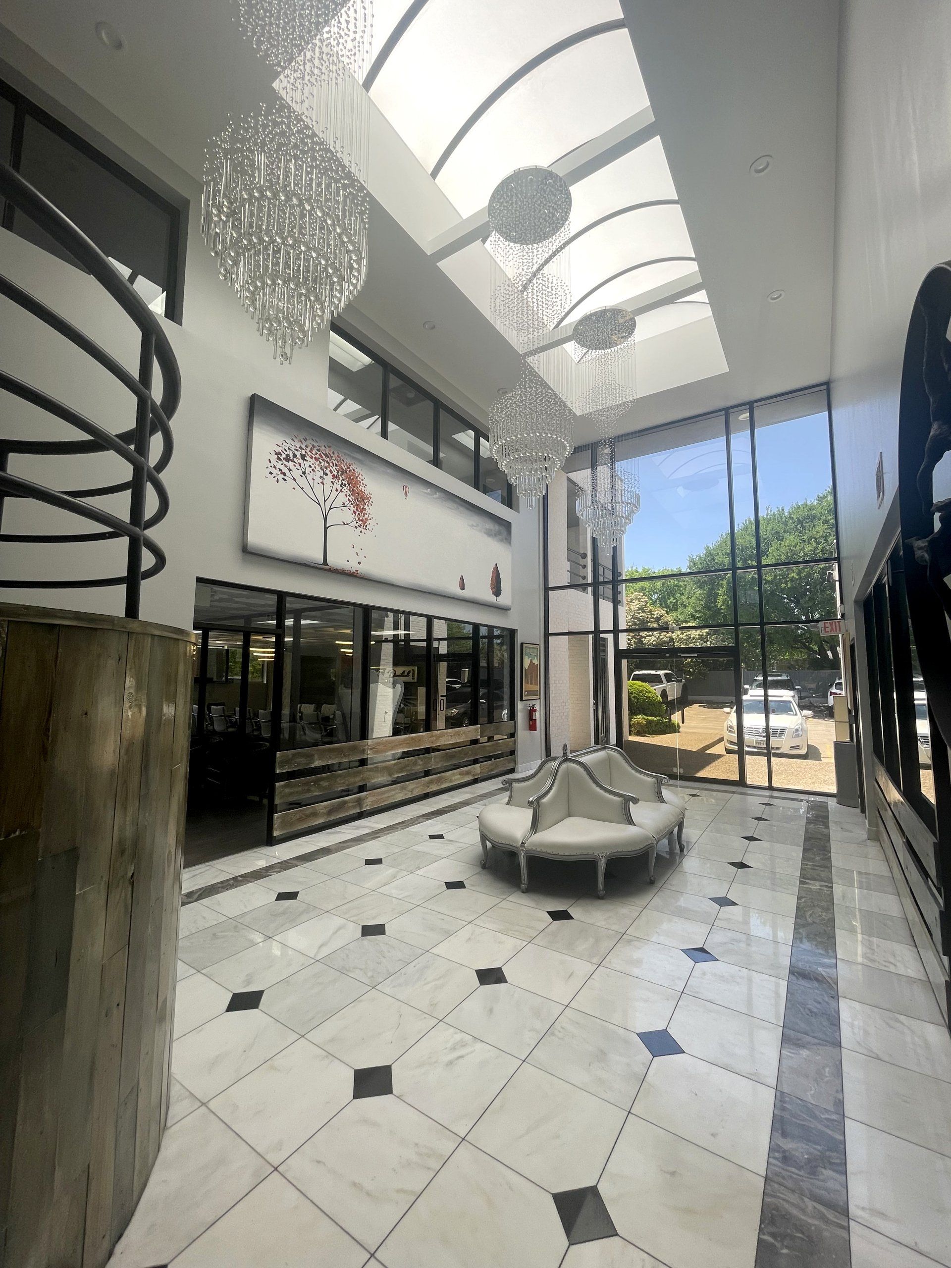 Modern covered courtyard with tiled floor, benches, glass storefronts, and skylit ceiling leading to outdoor trees.