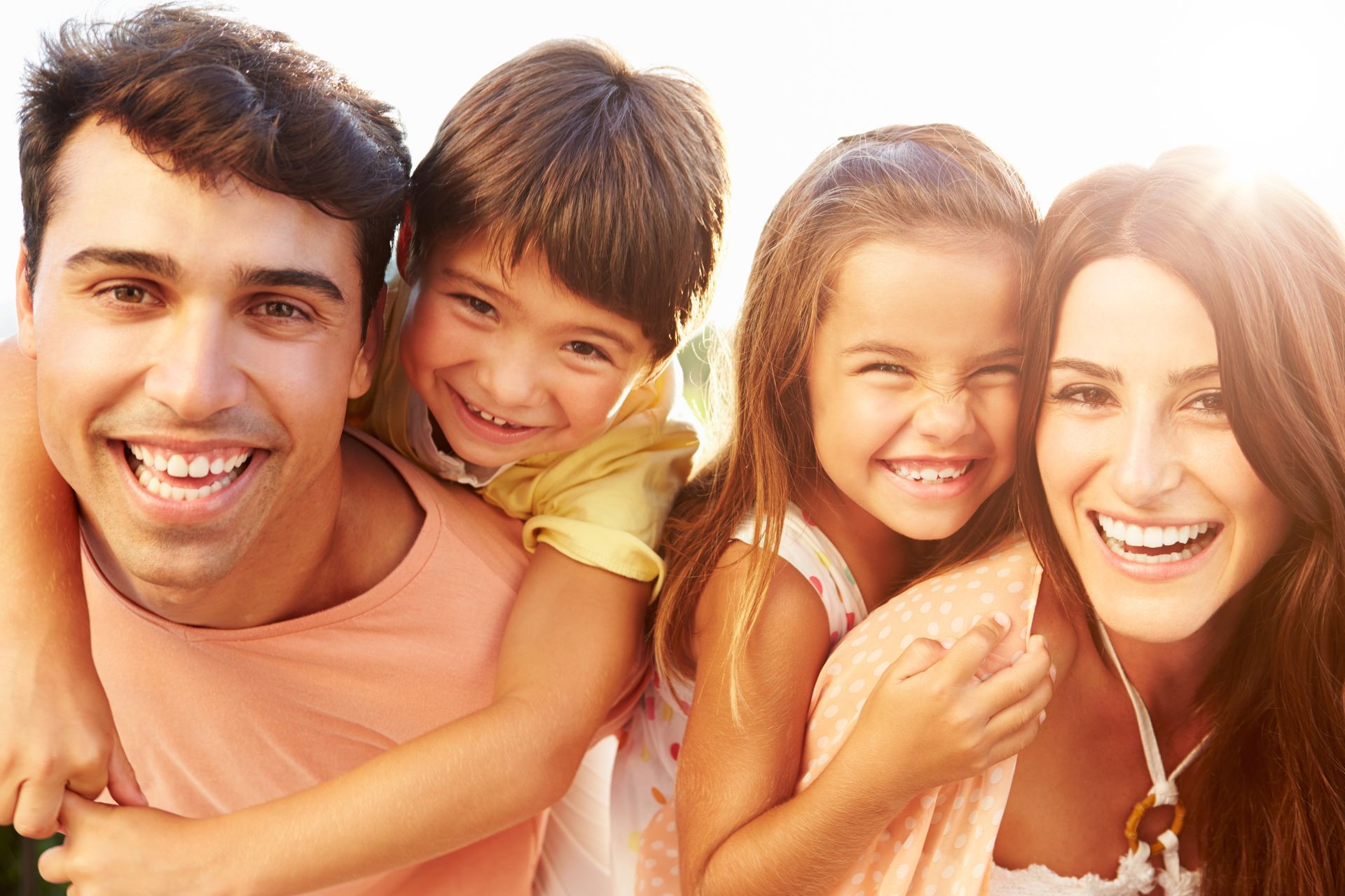 Smiling family outdoors in warm sunlight, two adults and two children posing closely together.