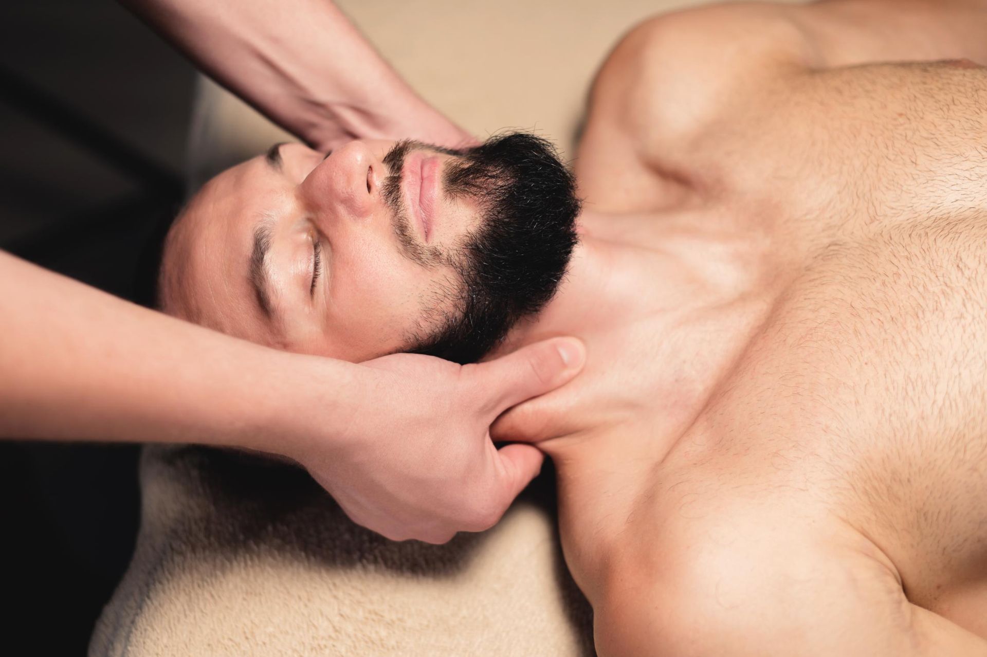 Man receiving a neck massage on a treatment table, eyes closed, with hands supporting his head