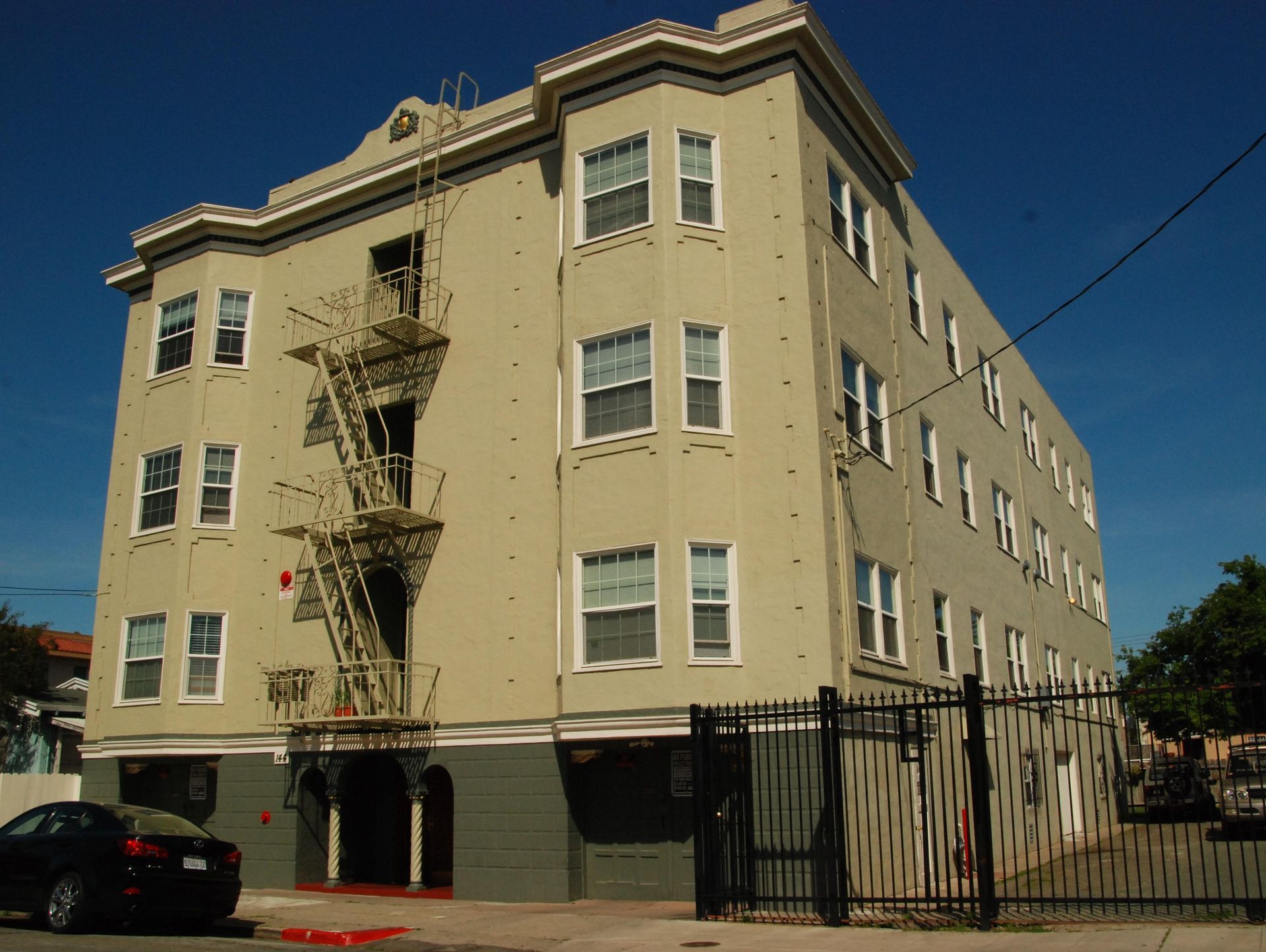 Beige four-story apartment building with fire escapes behind a black fence on a sunny street