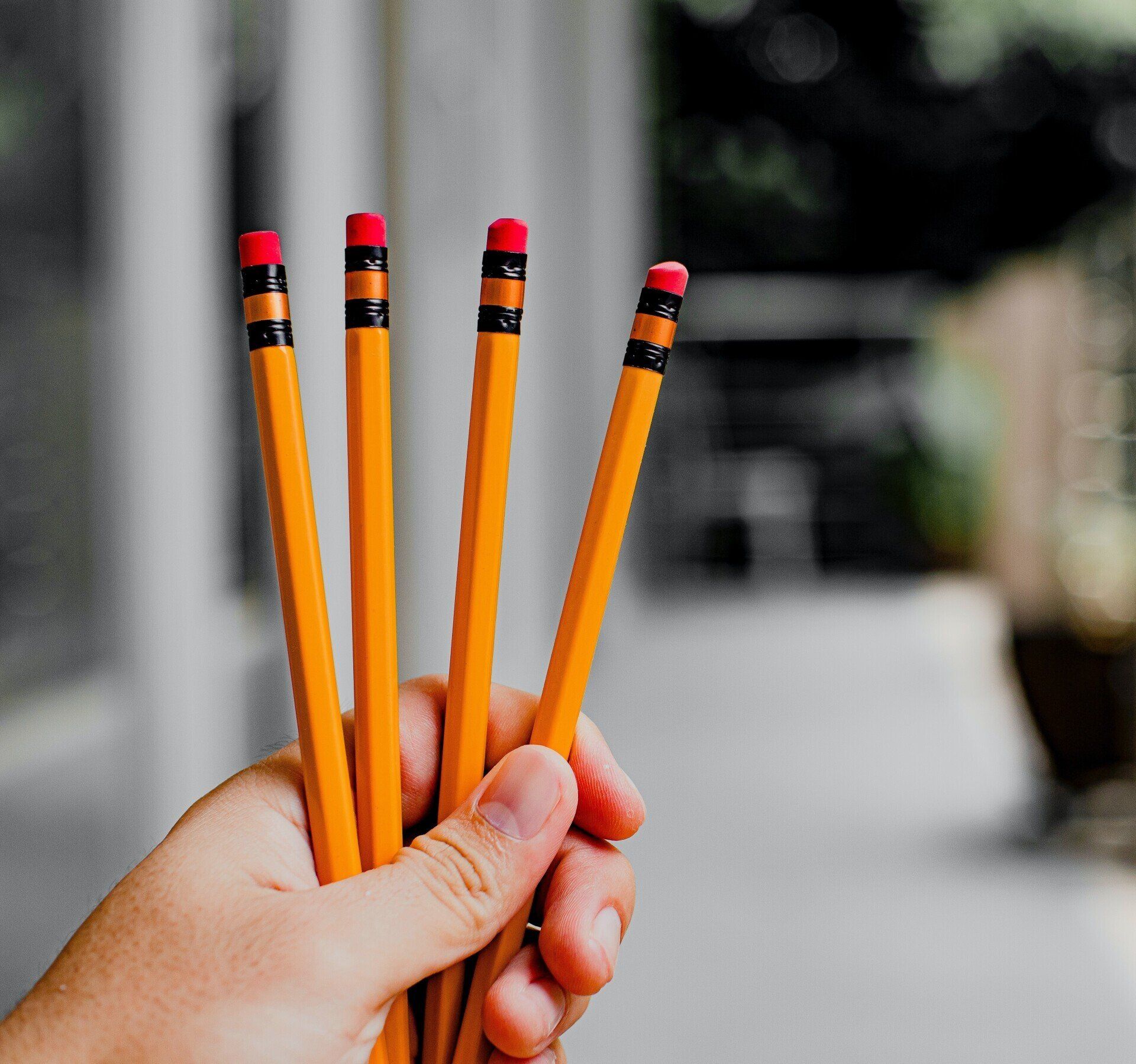 Hand holding four orange pencils with red erasers against a blurred outdoor background