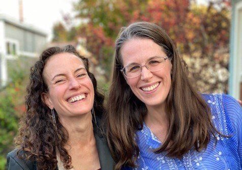 Two smiling women standing outdoors, one in a black top and one in a blue blouse, with autumn foliage behind them