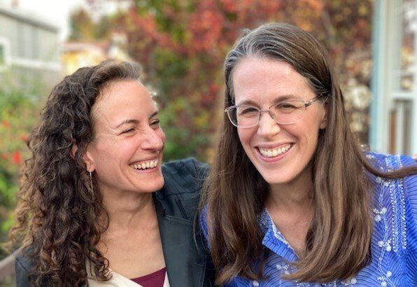 Two smiling women standing close together outdoors, one looking at the other, with a blurred garden background