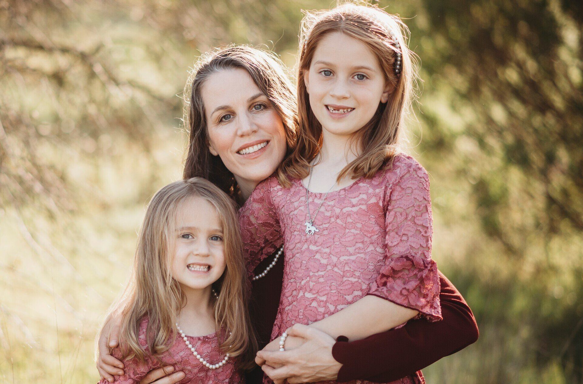 Two smiling children and a woman in matching pink tops standing outdoors in soft sunlight