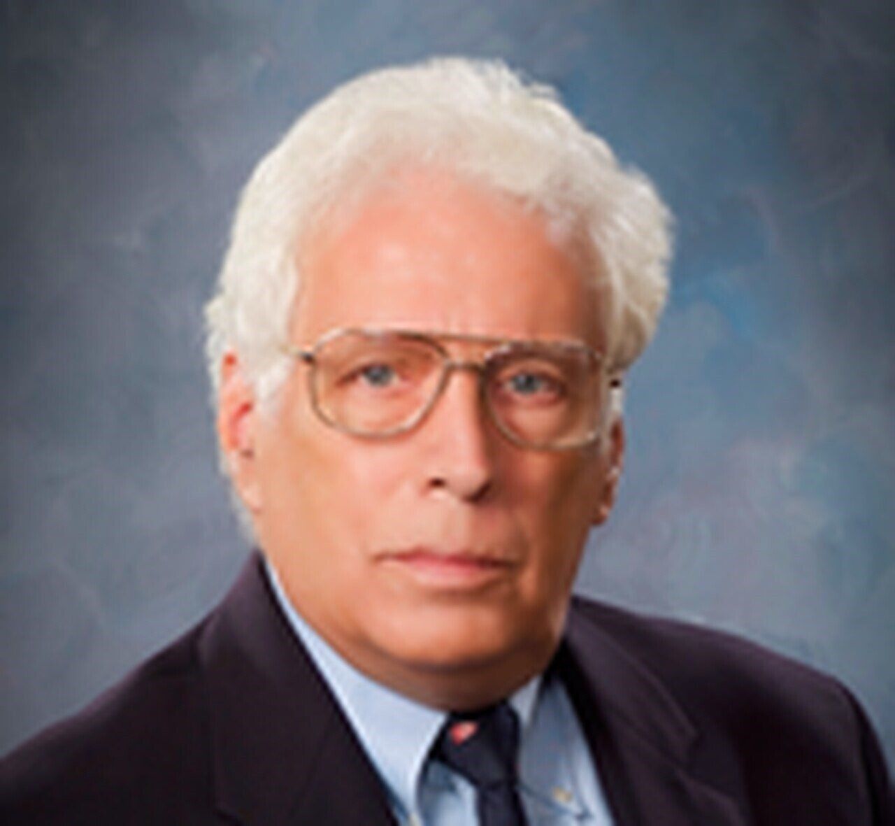 Portrait of a man in glasses wearing a dark suit and tie against a blue-gray studio backdrop