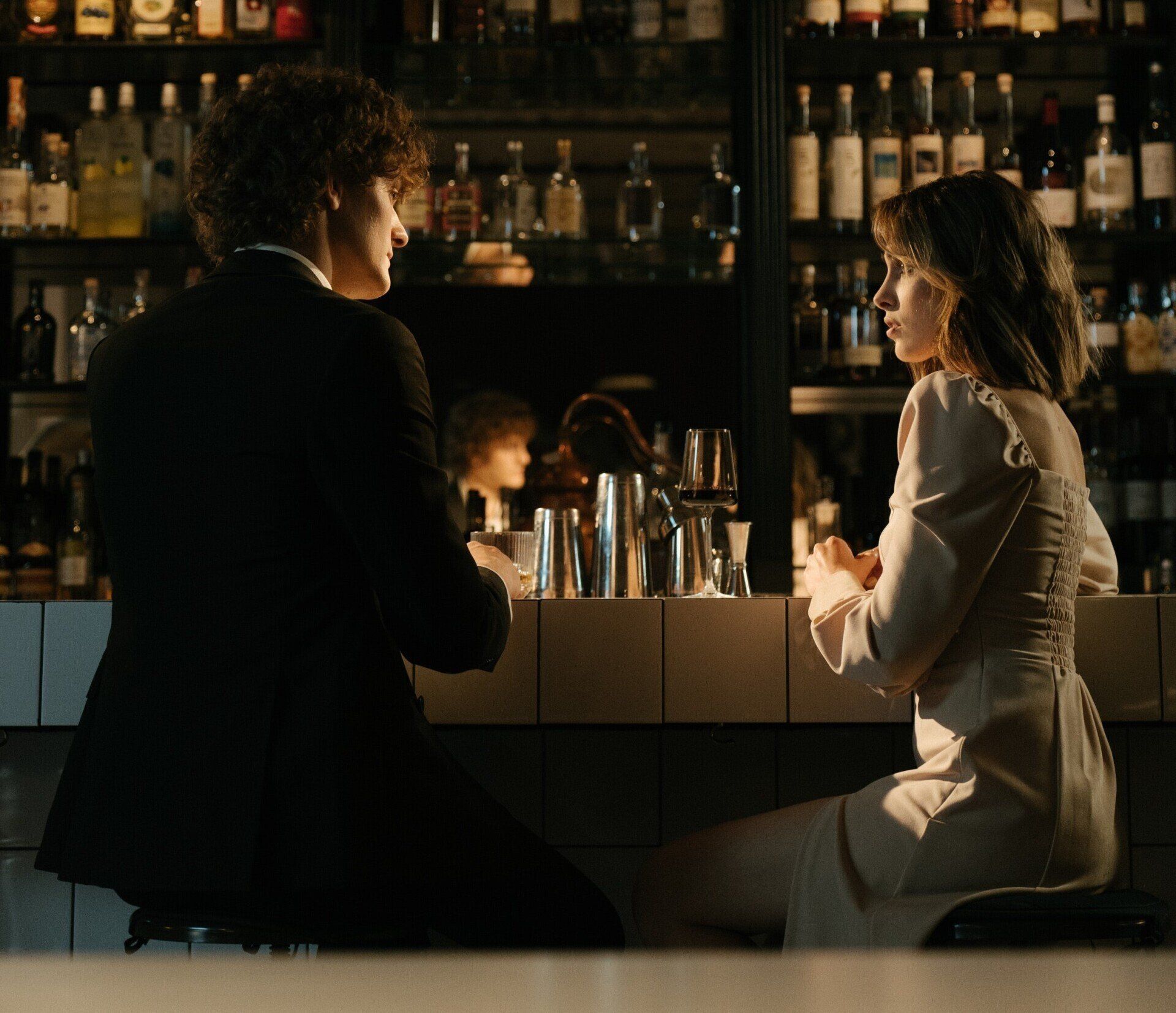 Two people talking at a dimly lit bar with shelves of bottles in the background
