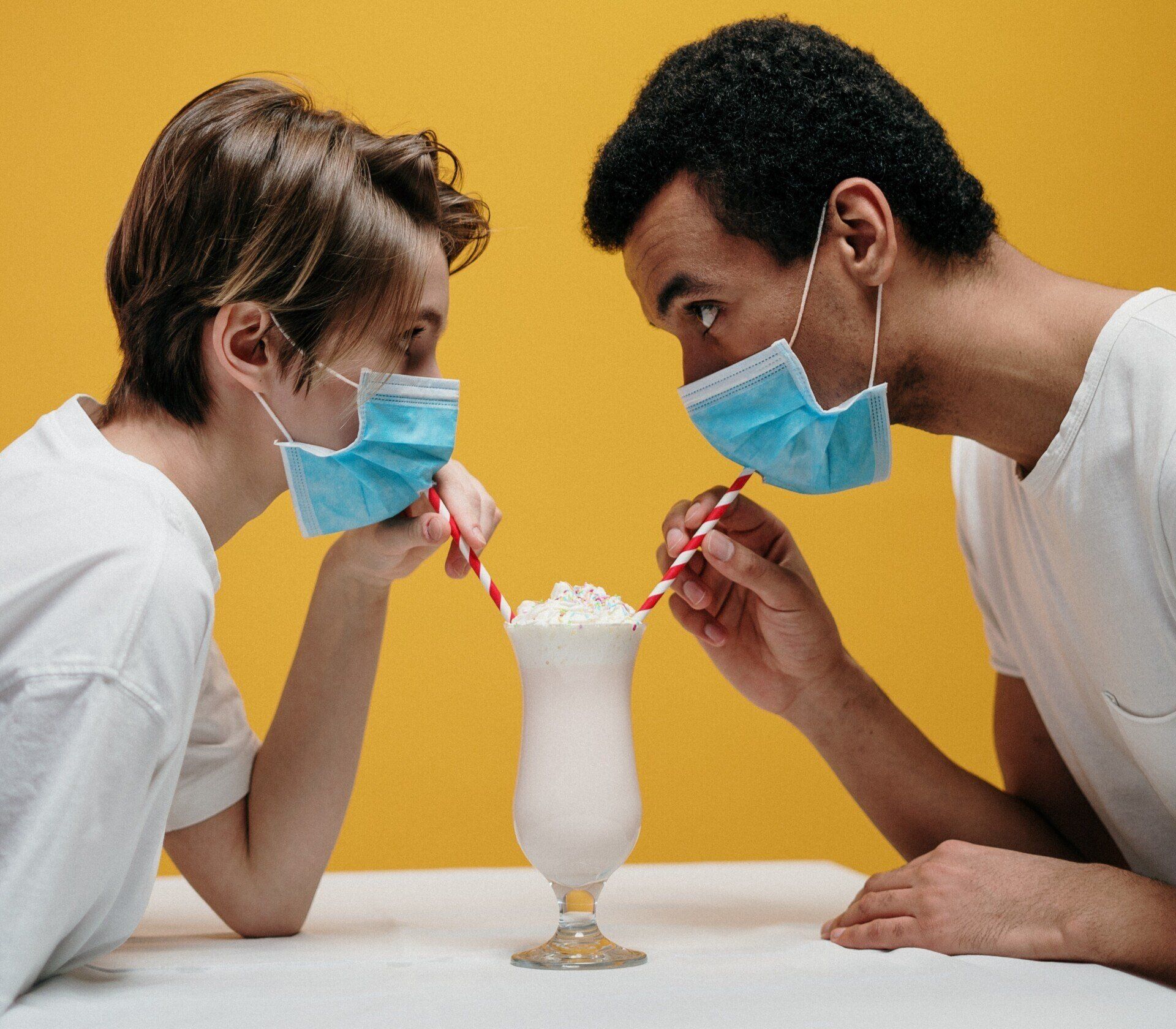 Two masked people lean toward a milkshake, each holding a red-and-white striped straw.