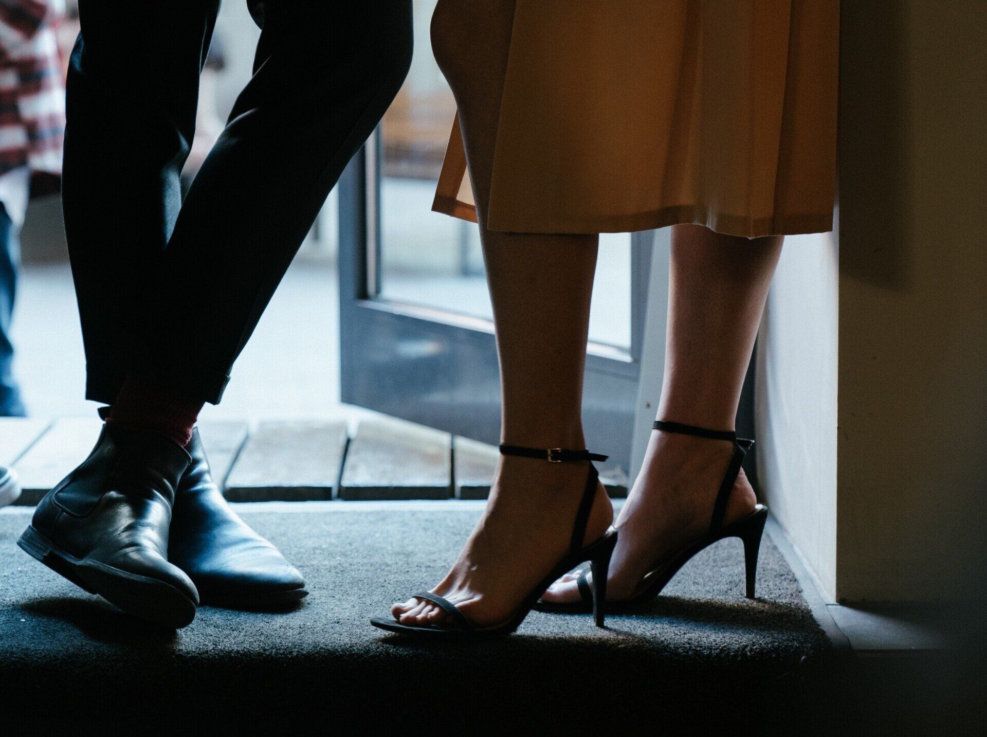 Two people standing by a window, one in black shoes and pants, the other in heels and a tan skirt.