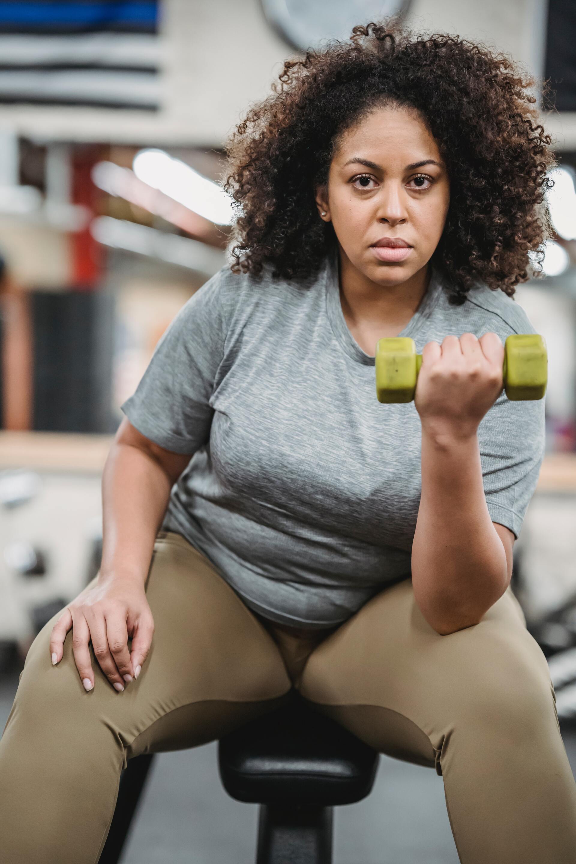 Woman in gray shirt lifting a green dumbbell while seated on a gym bench