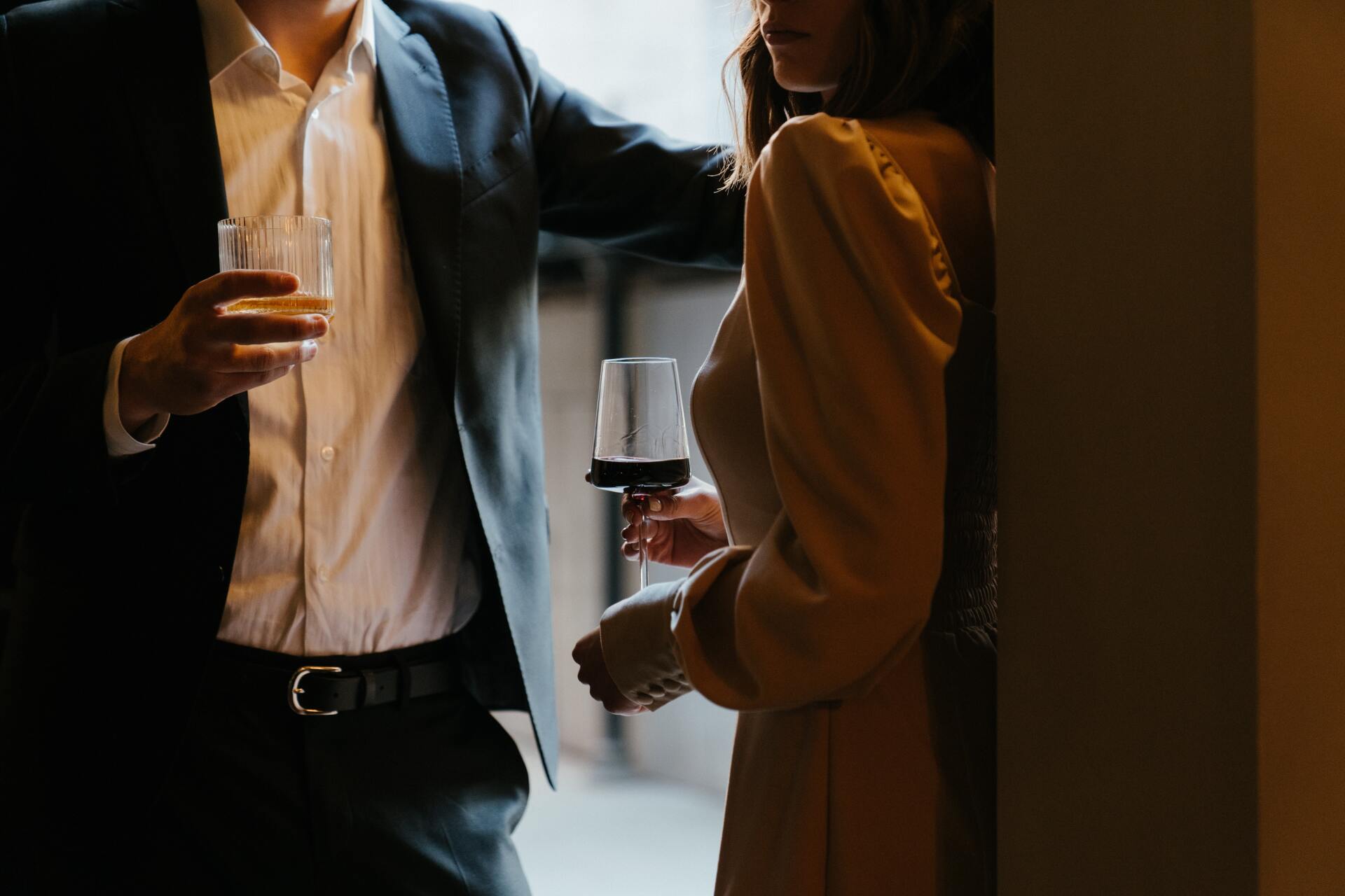 Two people talking near a doorway, one holding a drink in a dimly lit room