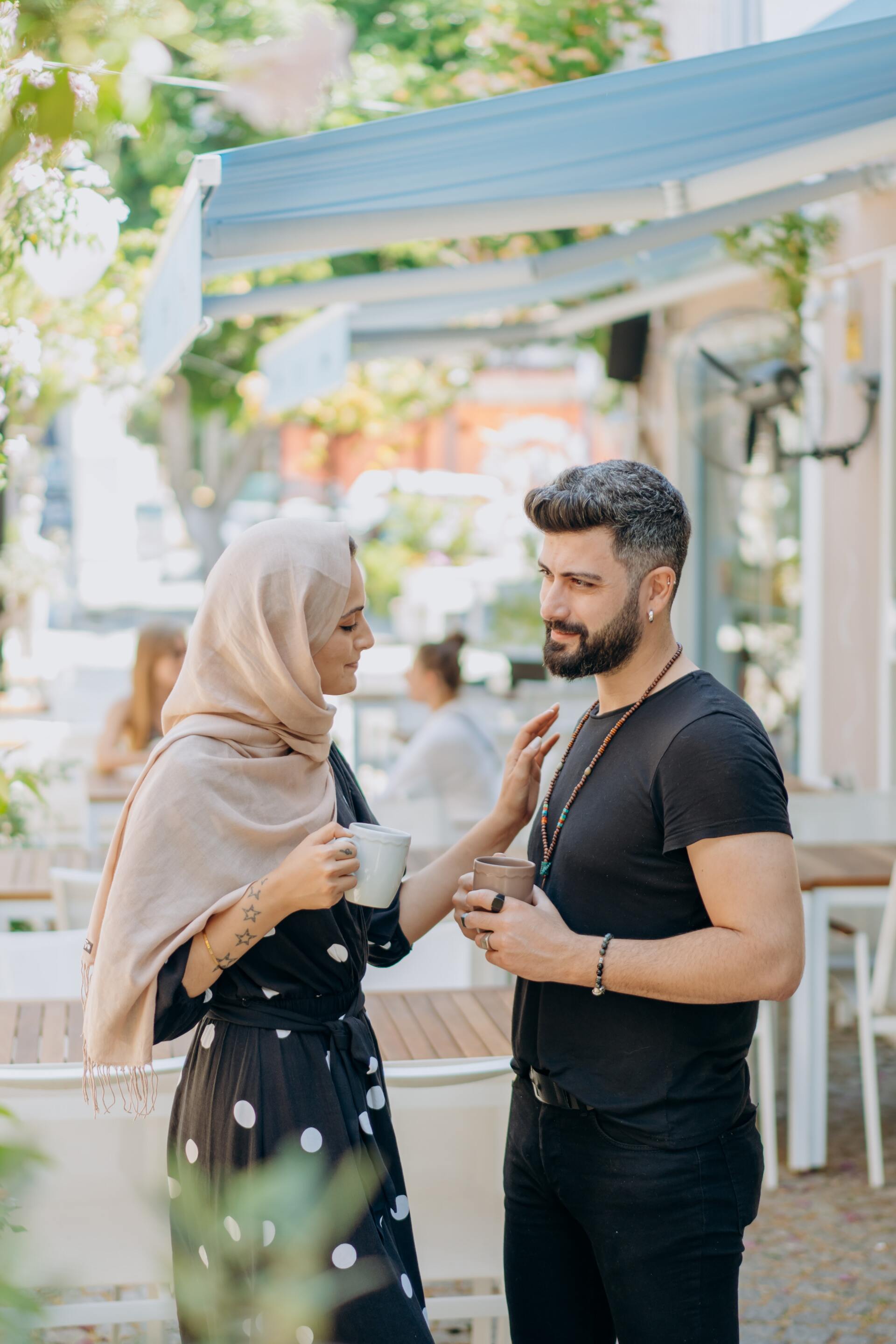 Two people talking under a white canopy at an outdoor café, holding drinks and smiling.