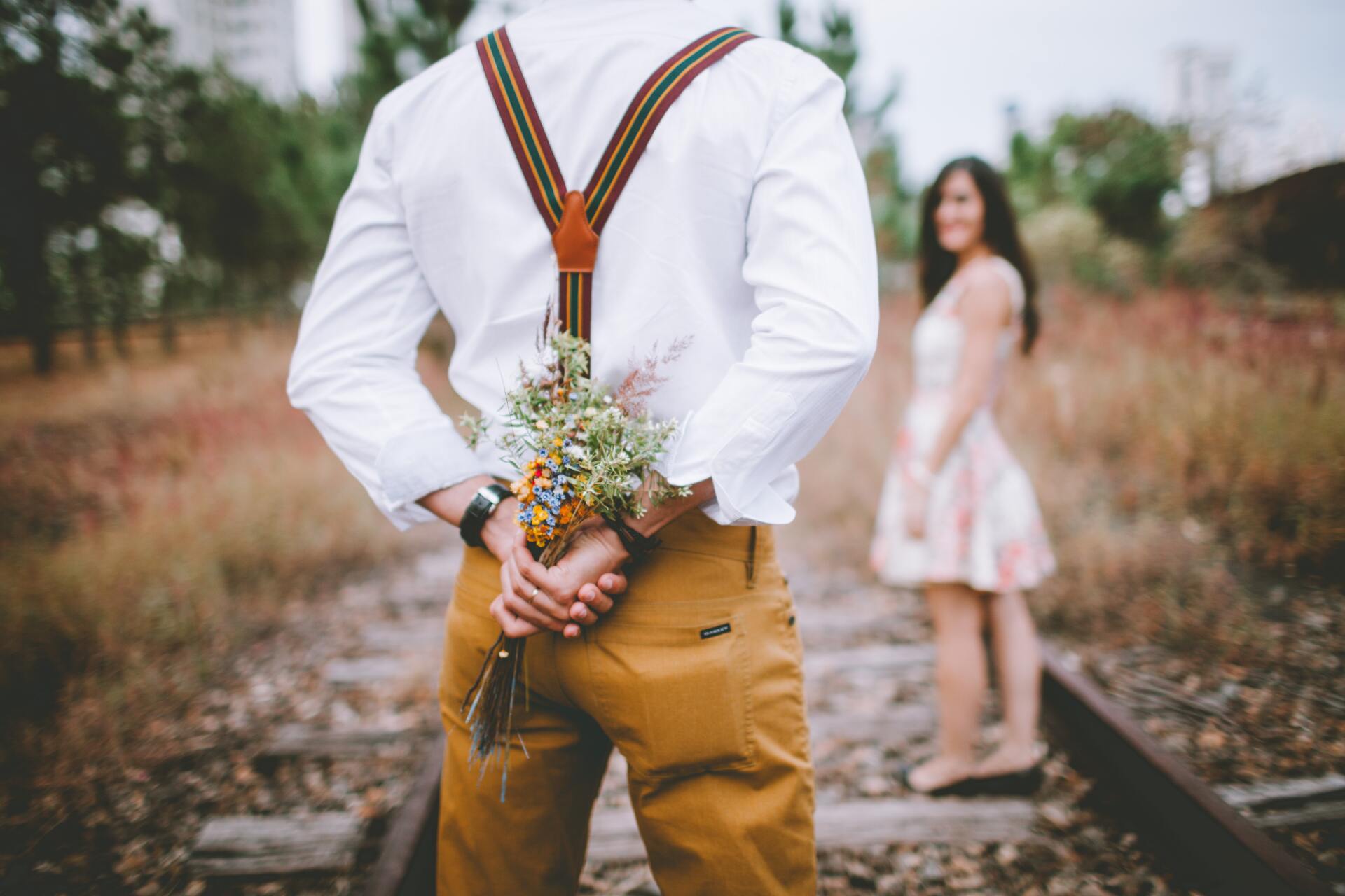 Man standing on railroad tracks holding flowers behind his back, with a woman in a white dress in the distance