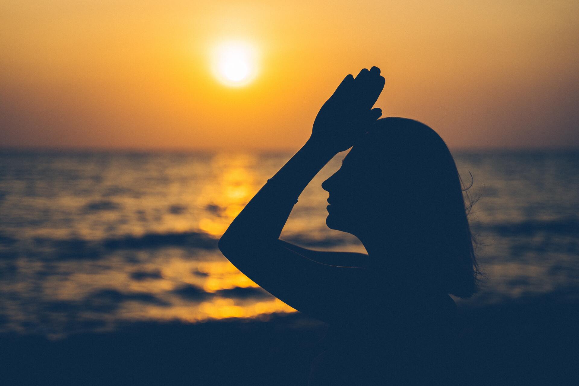 Silhouette of a person at sunset by the ocean, hand shielding their eyes as the sun sets over the water