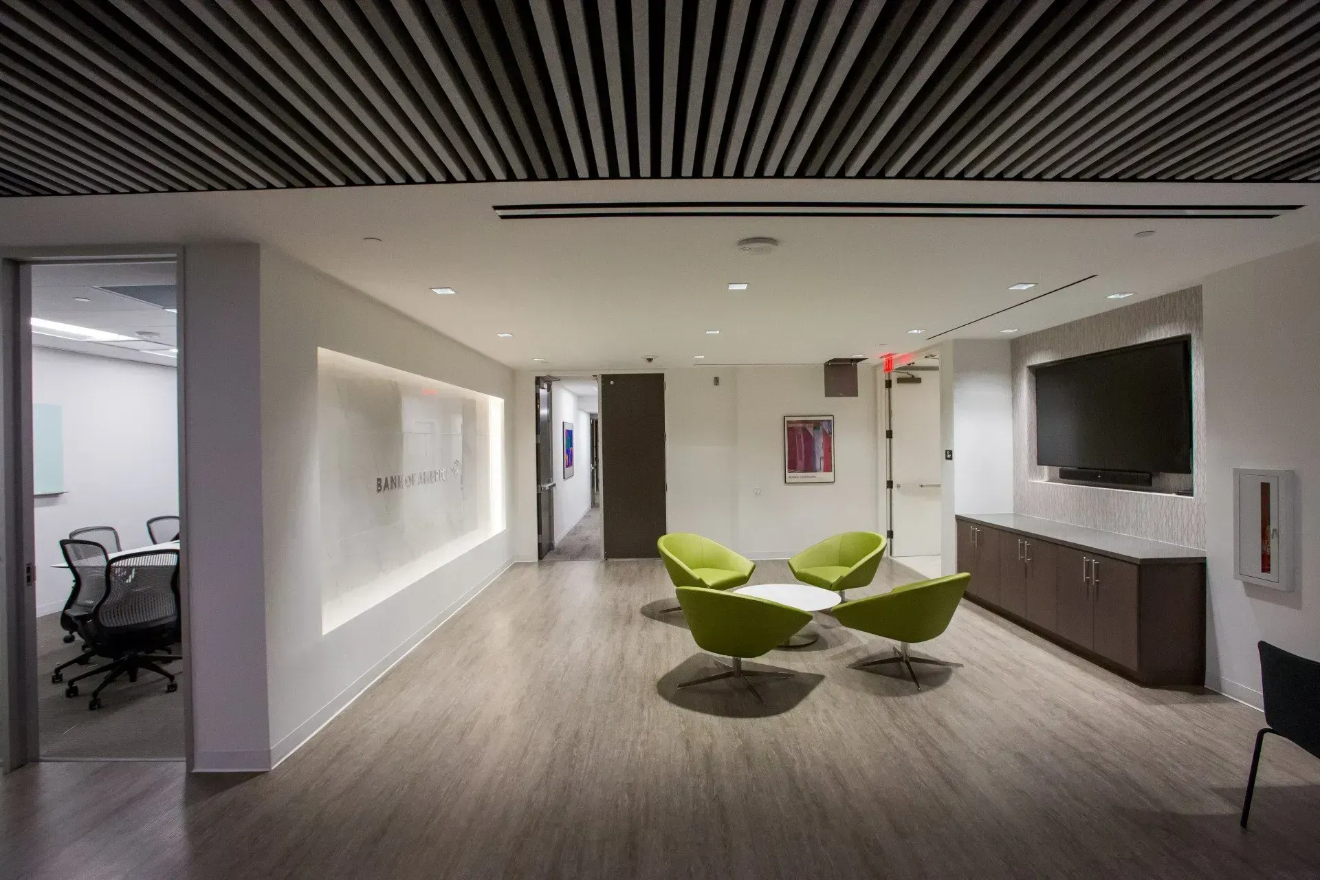 Modern office lounge with green chairs, wall-mounted TV, and conference room visible through a glass doorway