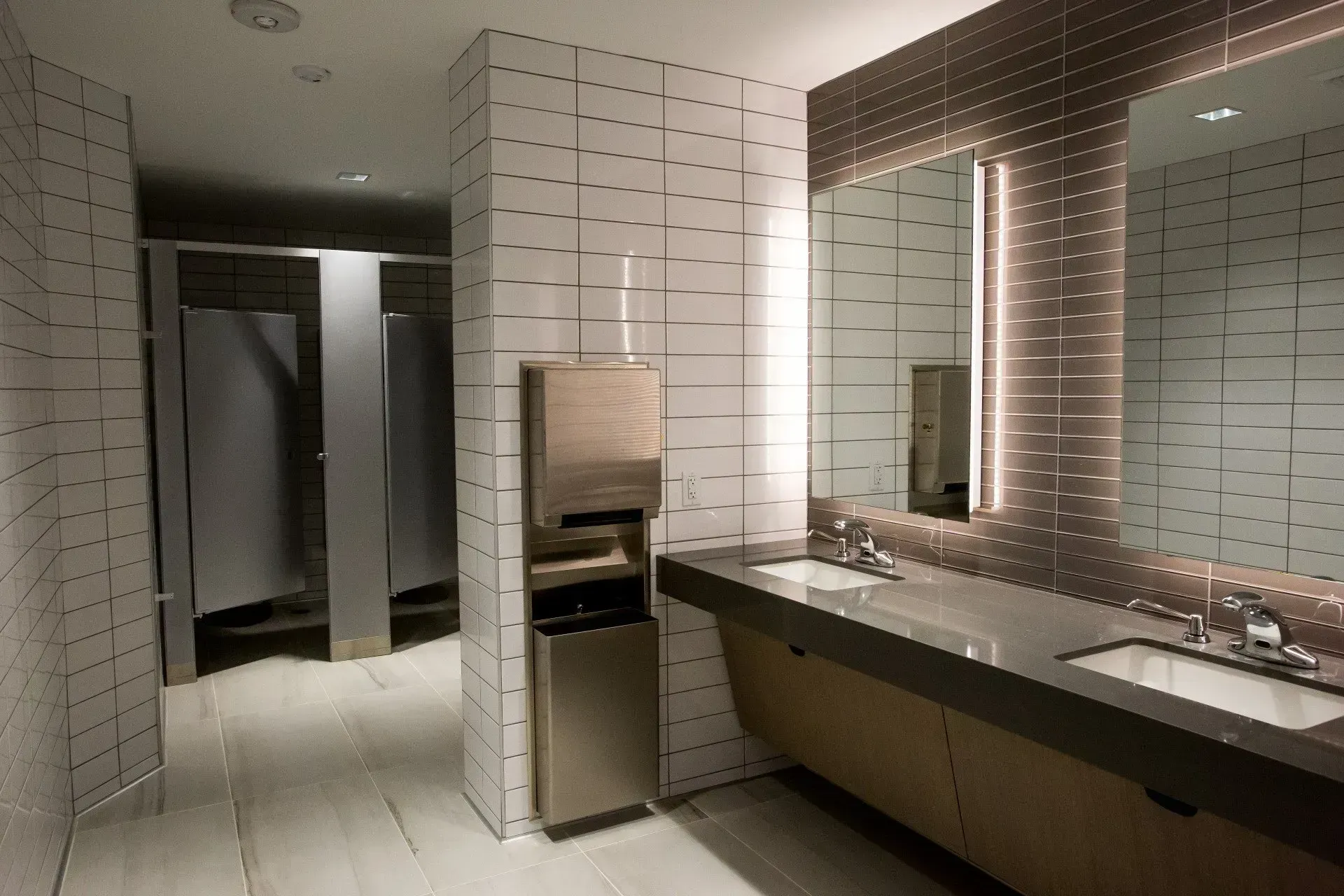 Public restroom with tiled walls, stalls, and long sinks beneath illuminated mirrors.