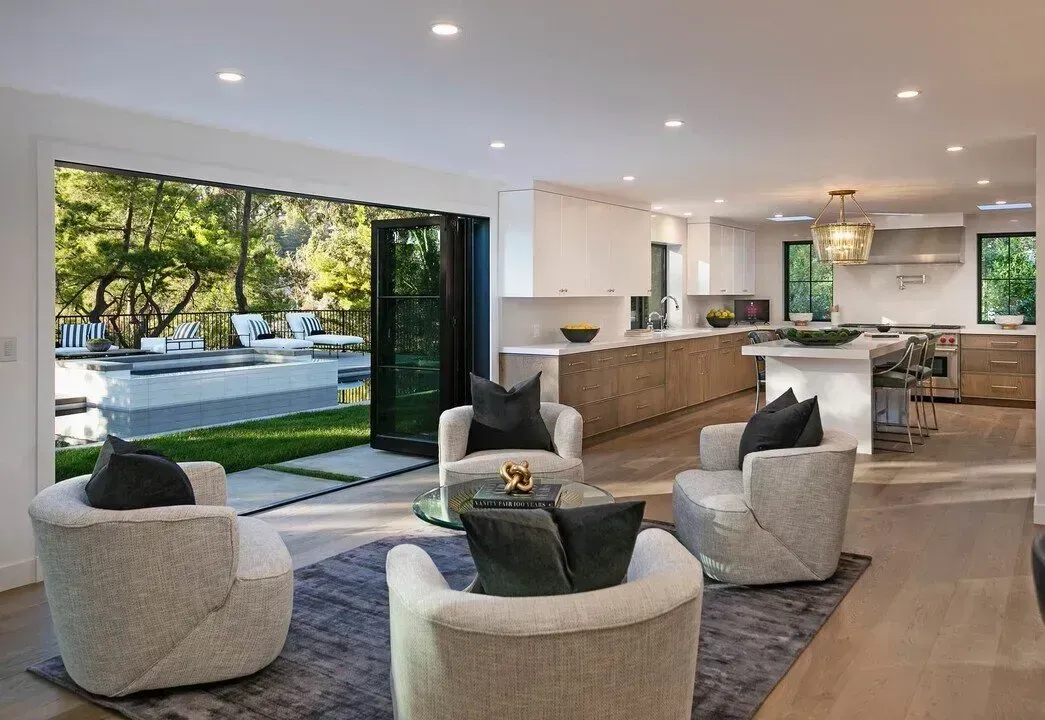 Modern open-plan living room with white chairs, gray rug, kitchen island, and large sliding doors to a sunny street view