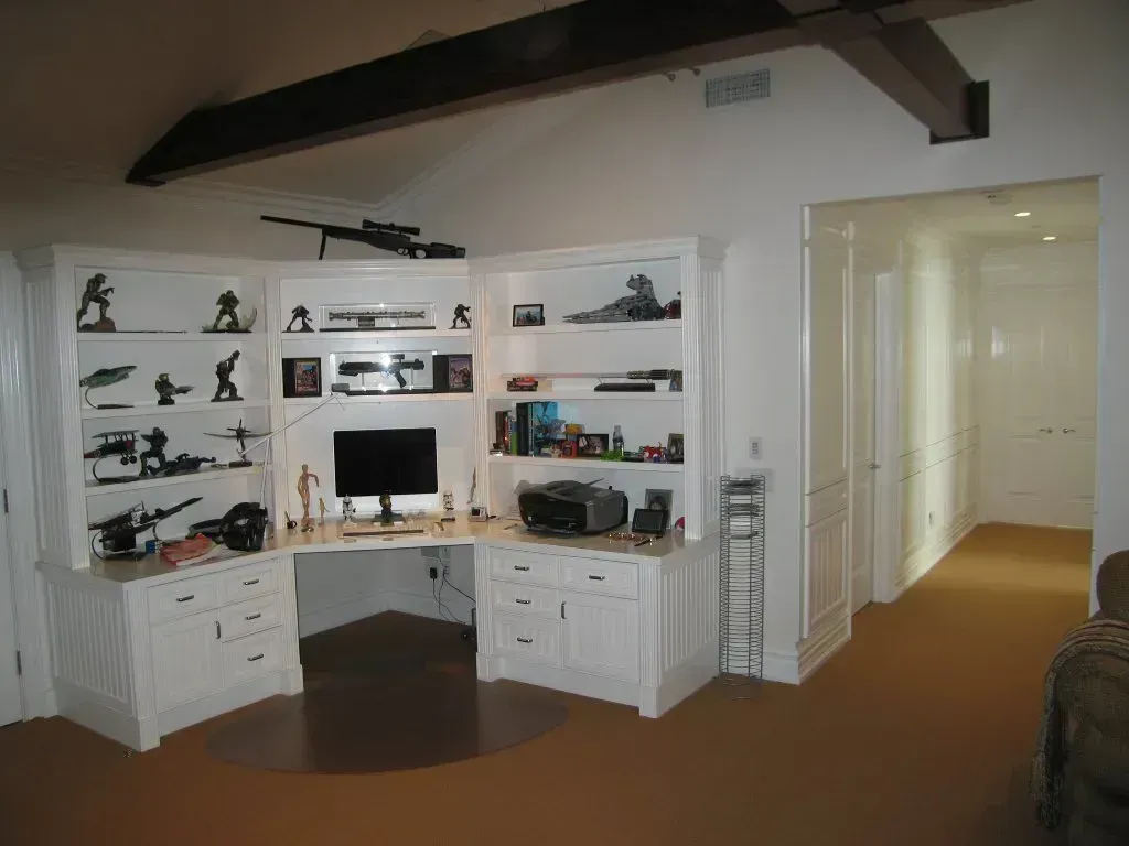 White corner home office with shelves, desk, monitor, and white cabinets in a room with brown carpet.