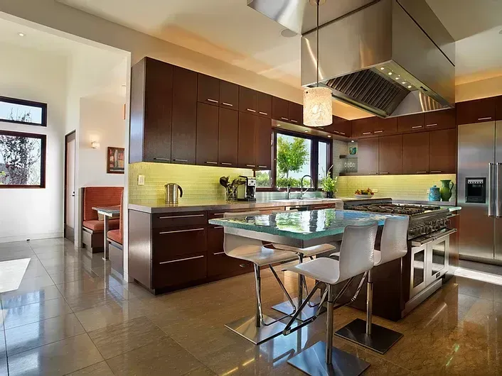 Modern kitchen with dark wood cabinets, glass island table, stainless hood, and large windows