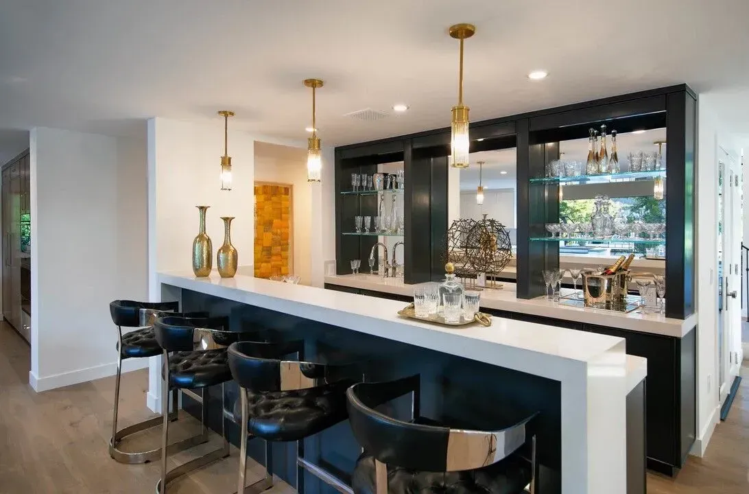 Modern home bar with black stools, white countertop, pendant lights, and glass-front cabinets