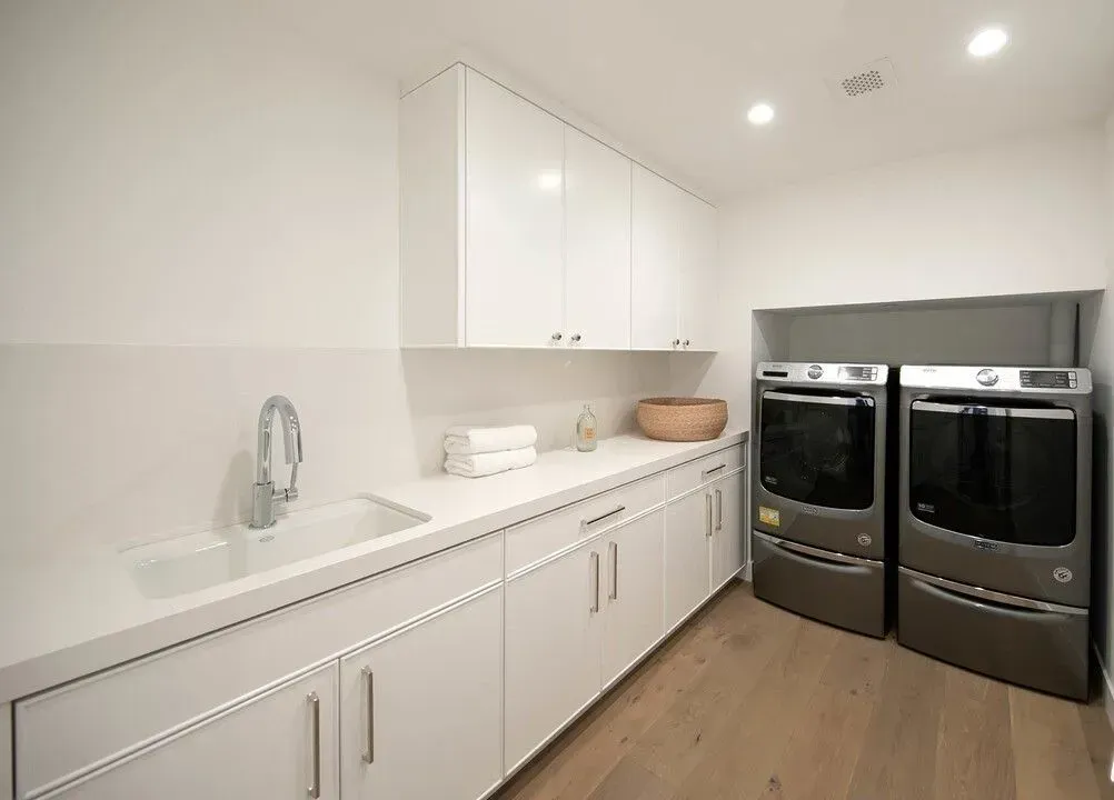 Bright white laundry room with long countertop, sink, cabinets, and two front-loading washer-dryers.