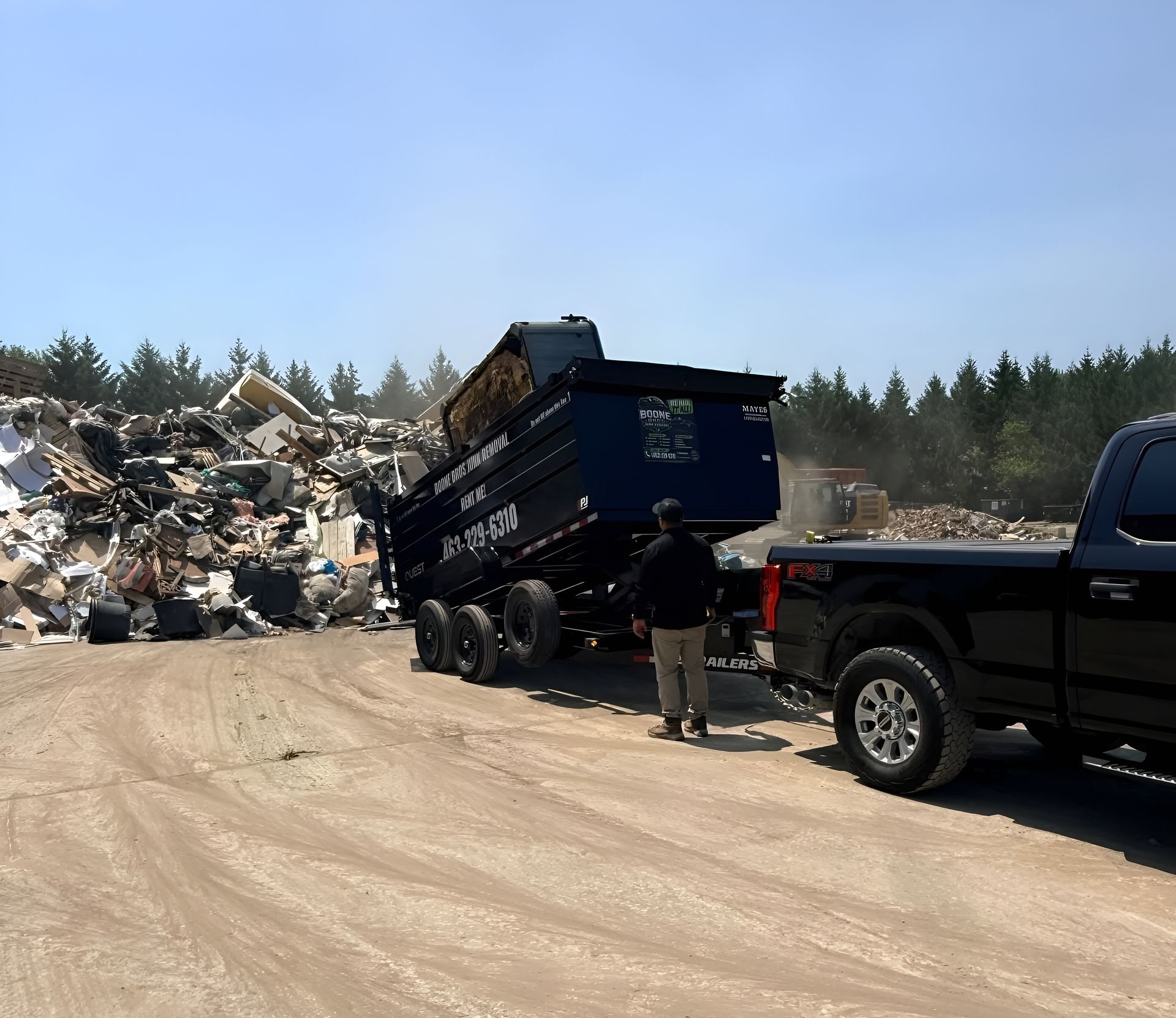 Blue dump trailer at a construction debris site beside a black pickup on a dusty road.