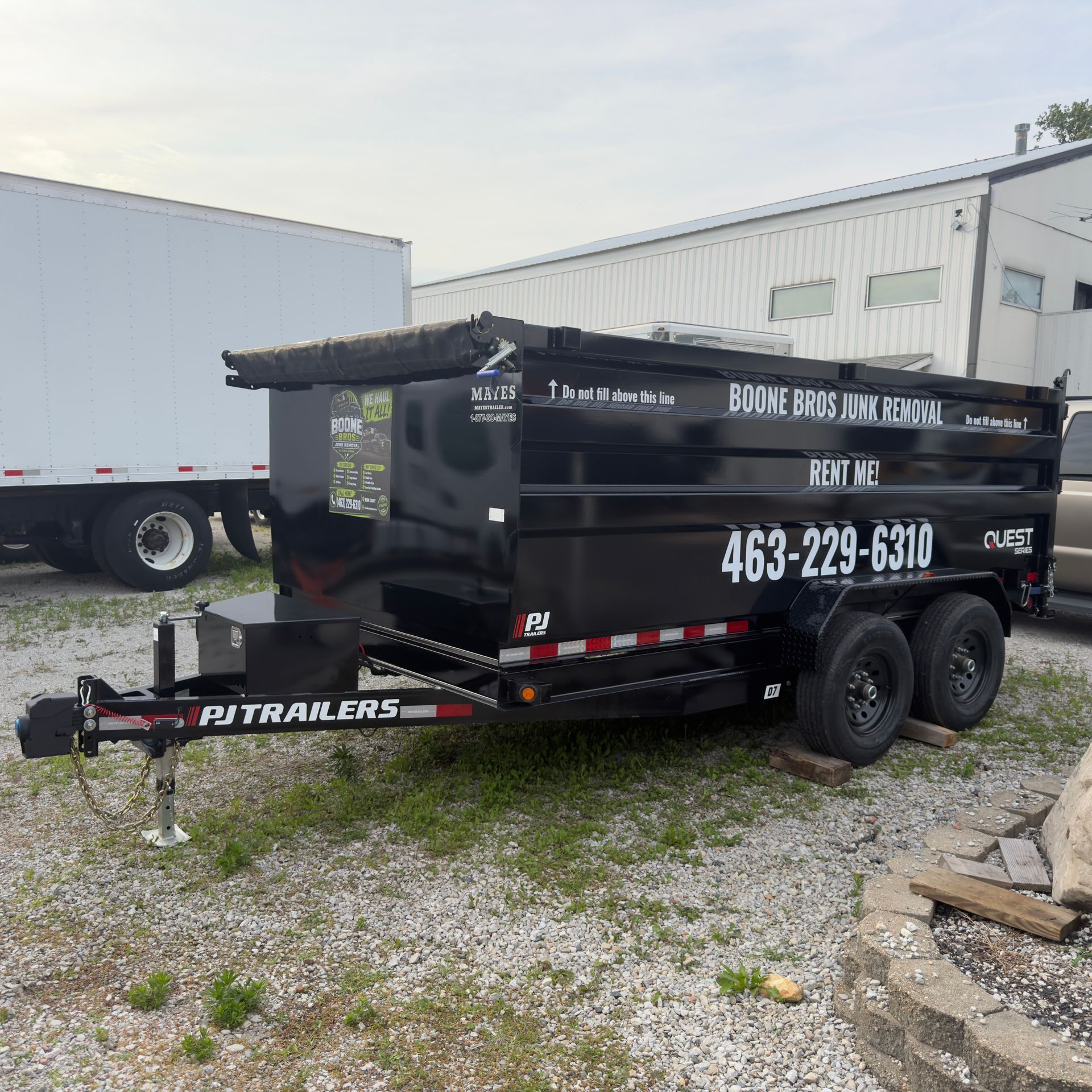 Black dump trailer with white lettering parked on gravel beside white warehouse buildings.