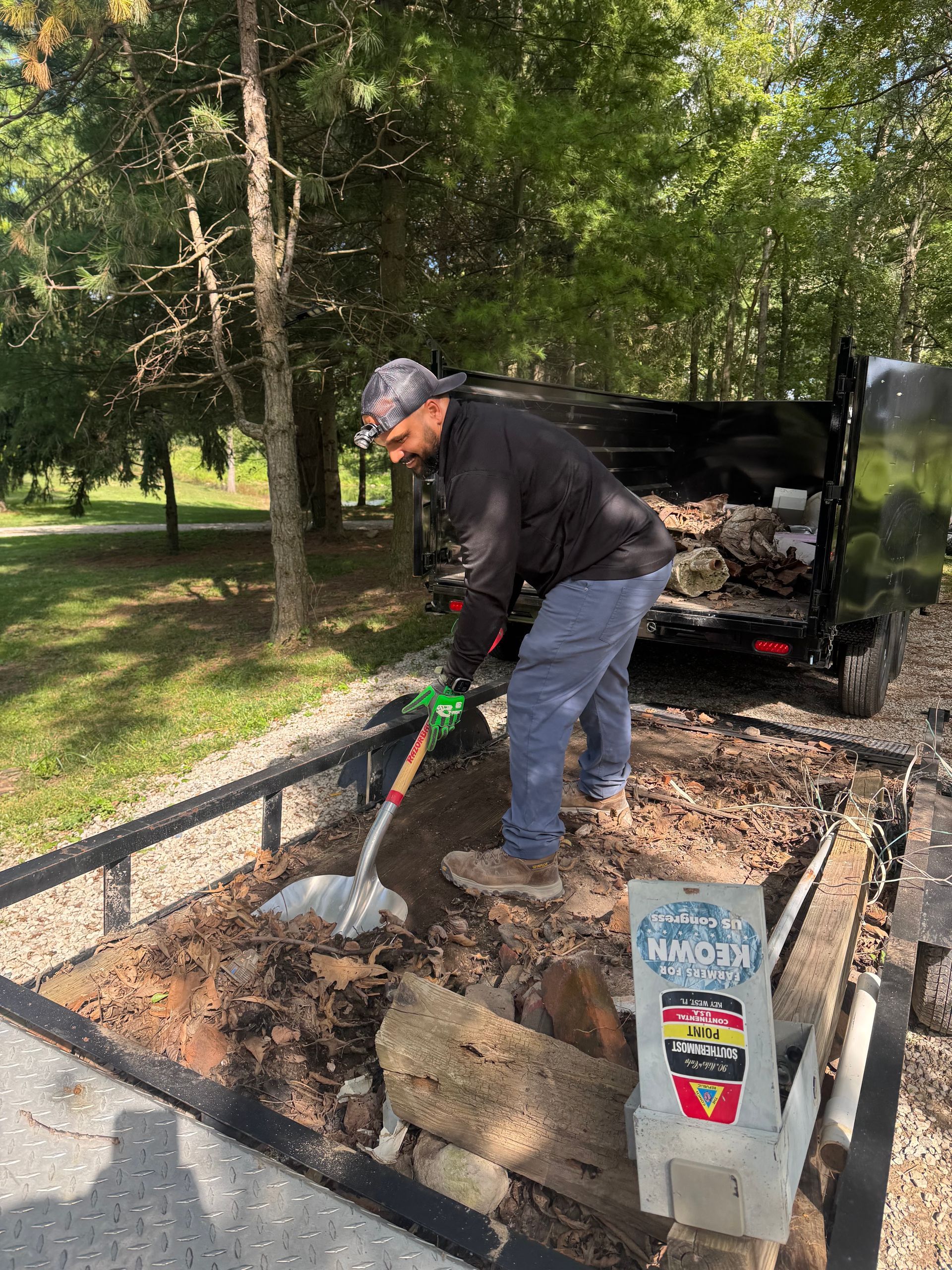 Worker shoveling dirt into a truck bed at a roadside work site with trees nearby