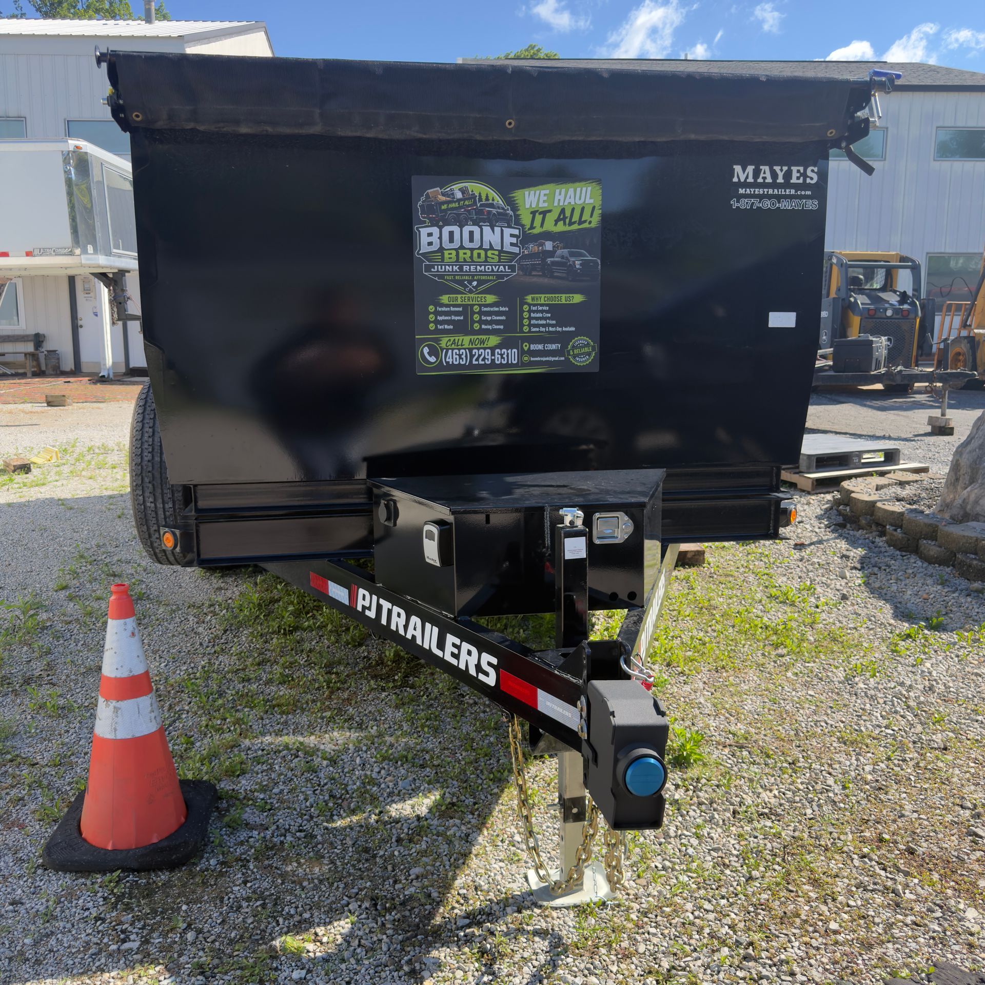 Black utility dump trailer with green sign, parked on gravel beside an orange cone.