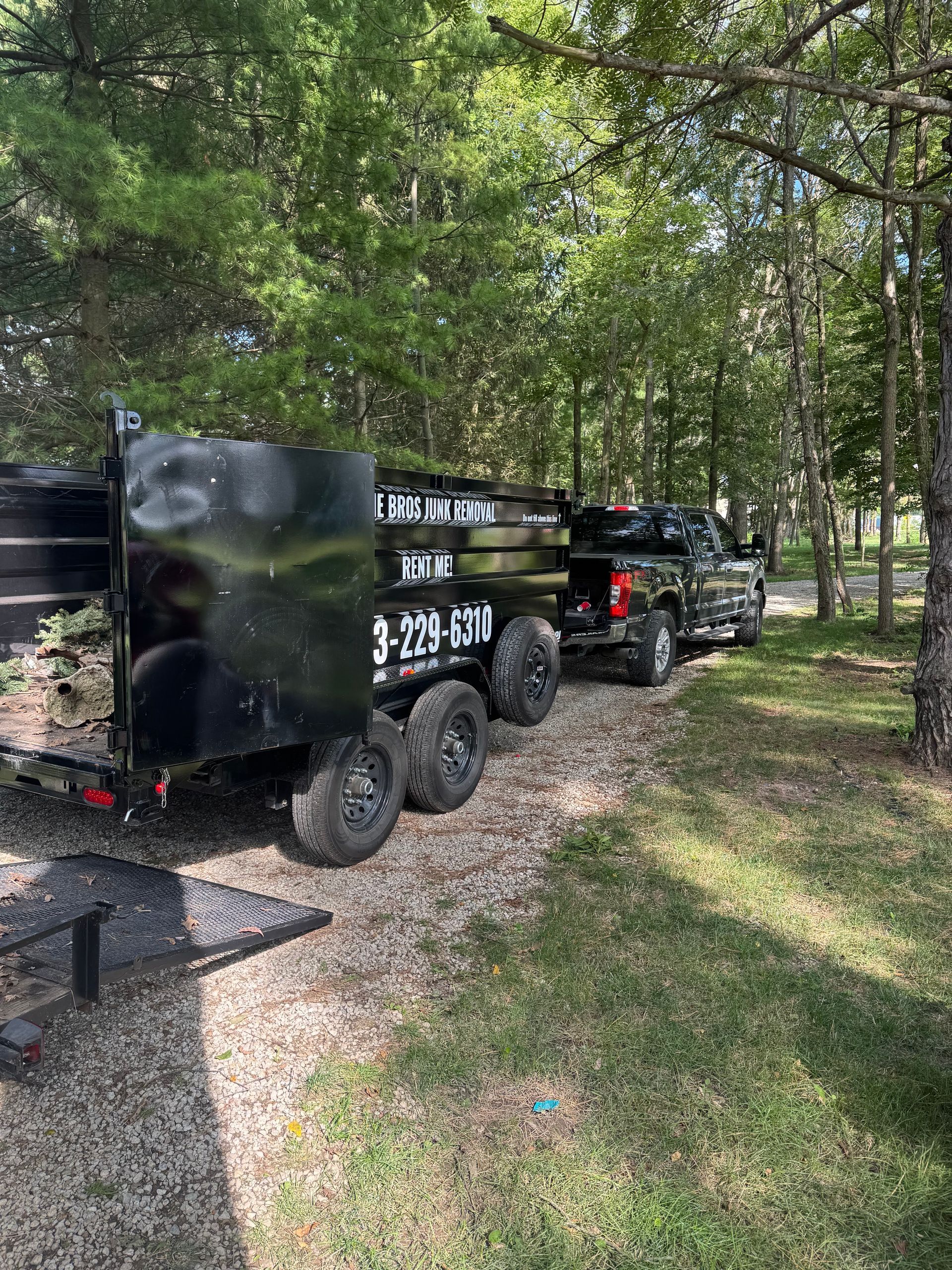 Black utility trailer and pickup truck parked on a gravel path in a wooded area.