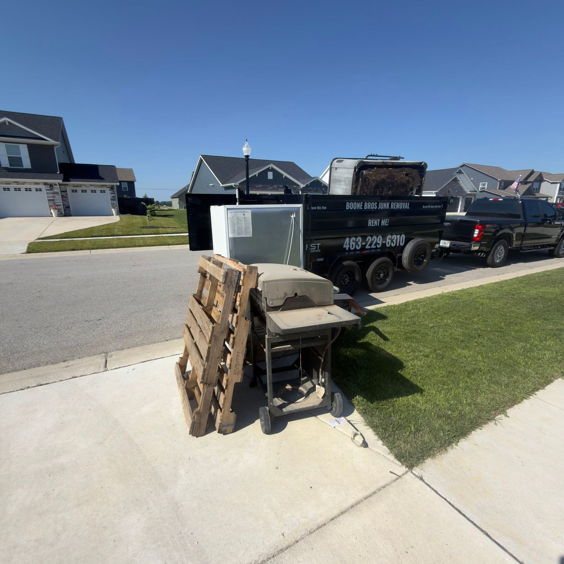 Black utility trailer with stacked boards and furniture on a suburban driveway beside a black pickup truck.