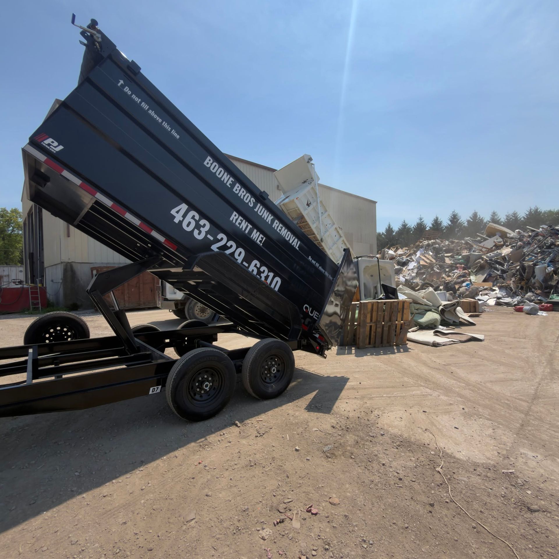 Black dump trailer tipped up at a scrapyard, with metal piles and dirt road under a clear sky.