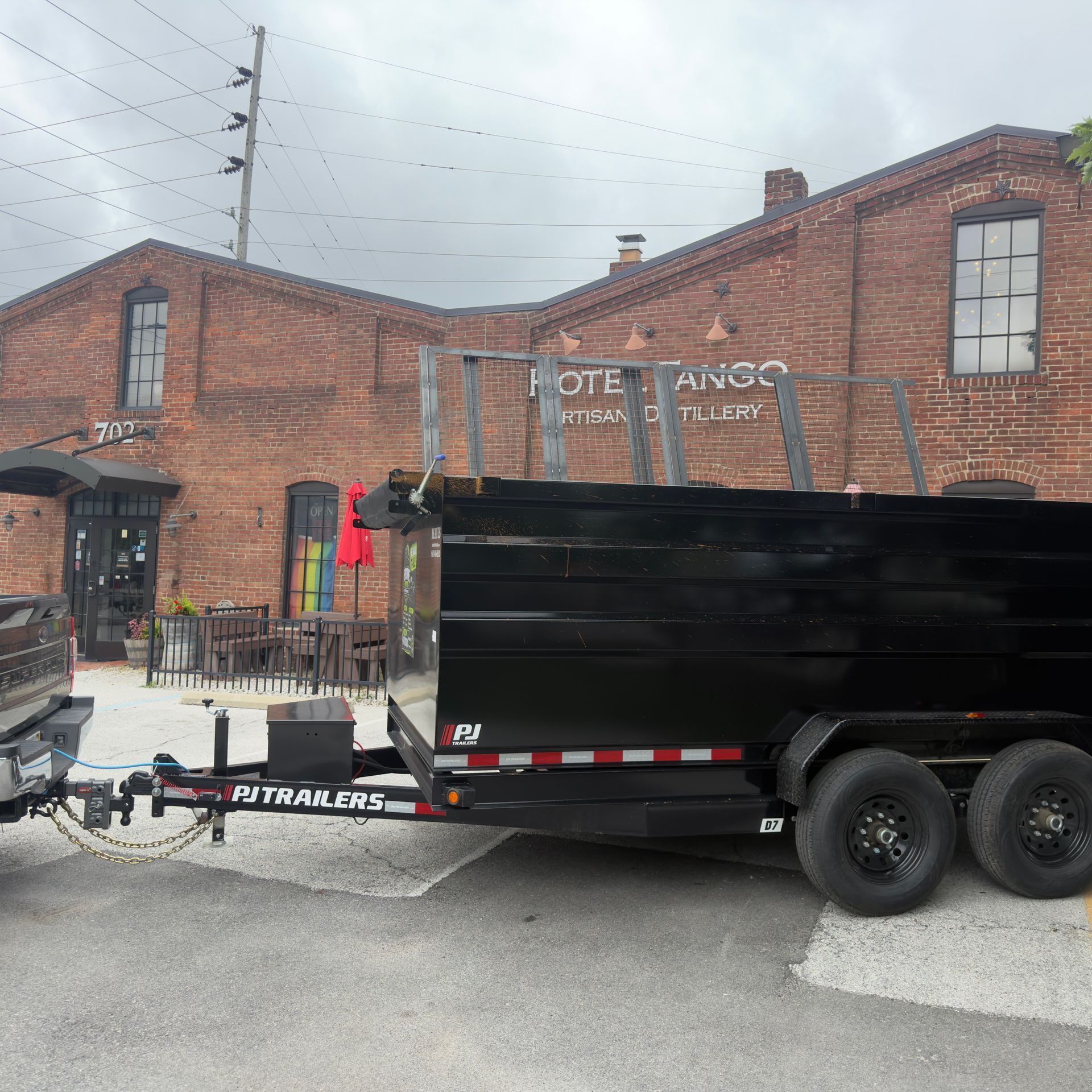 Black dump trailer parked beside a brick building with a fenced walkway and overcast sky