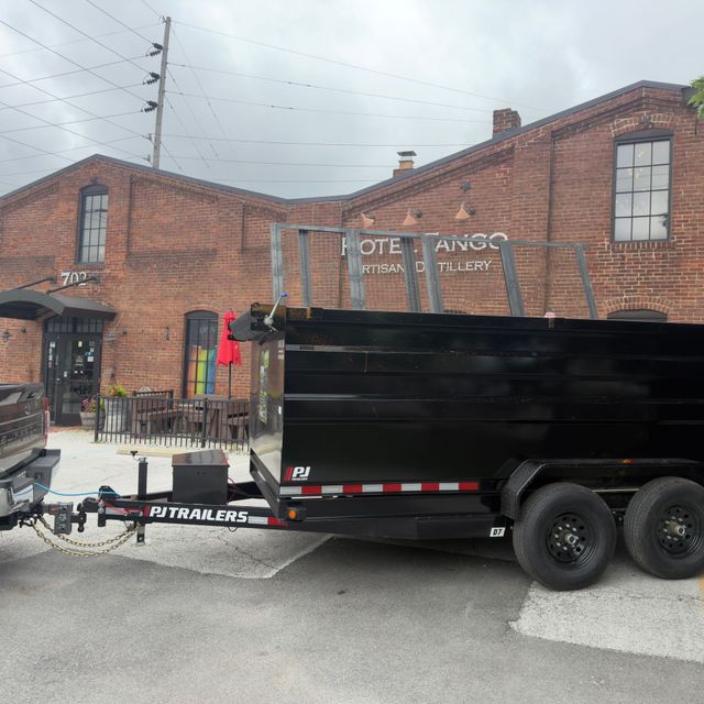Black dump trailer parked beside a brick building with a fenced walkway and overcast sky