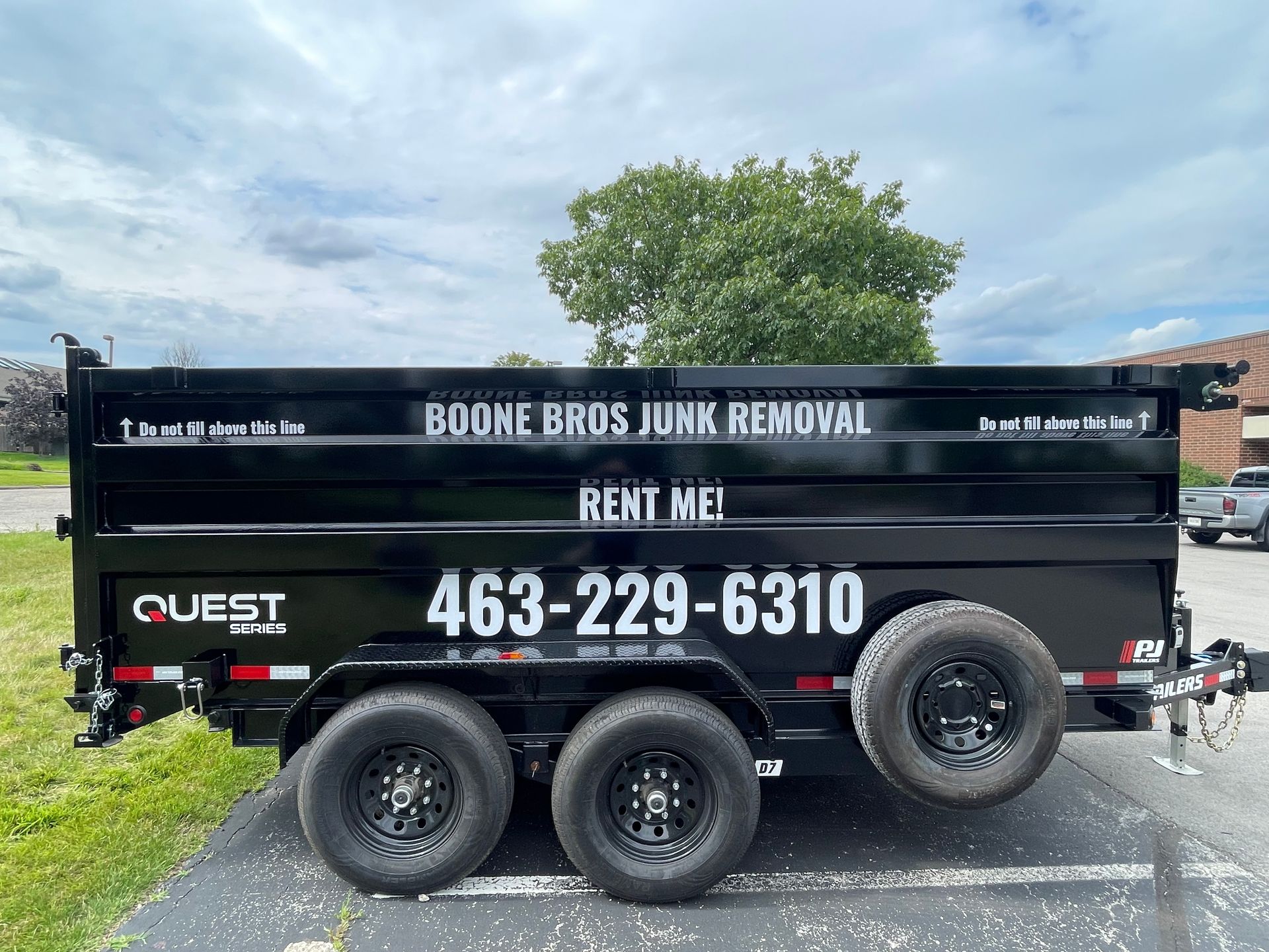 Black dump trailer advertising Boone Bros Junk Removal and rent me, parked on grass under cloudy sky