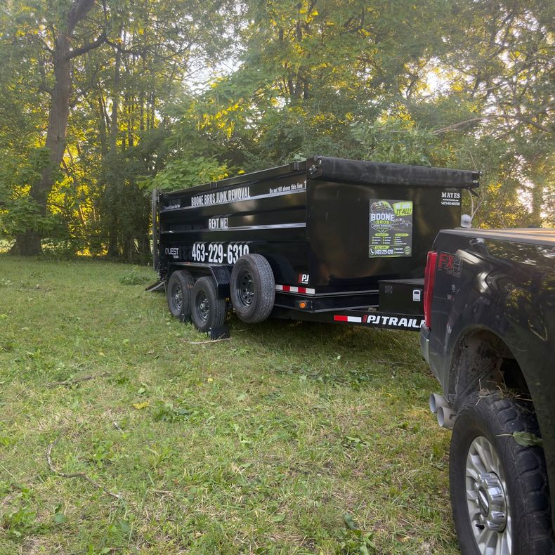 Black utility trailer with white lettering parked on grass beside a black truck near trees