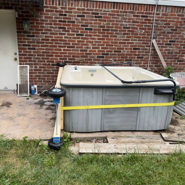 Above-ground hot tub in a backyard, with a brick wall and lawn nearby; yellow strap secures it.