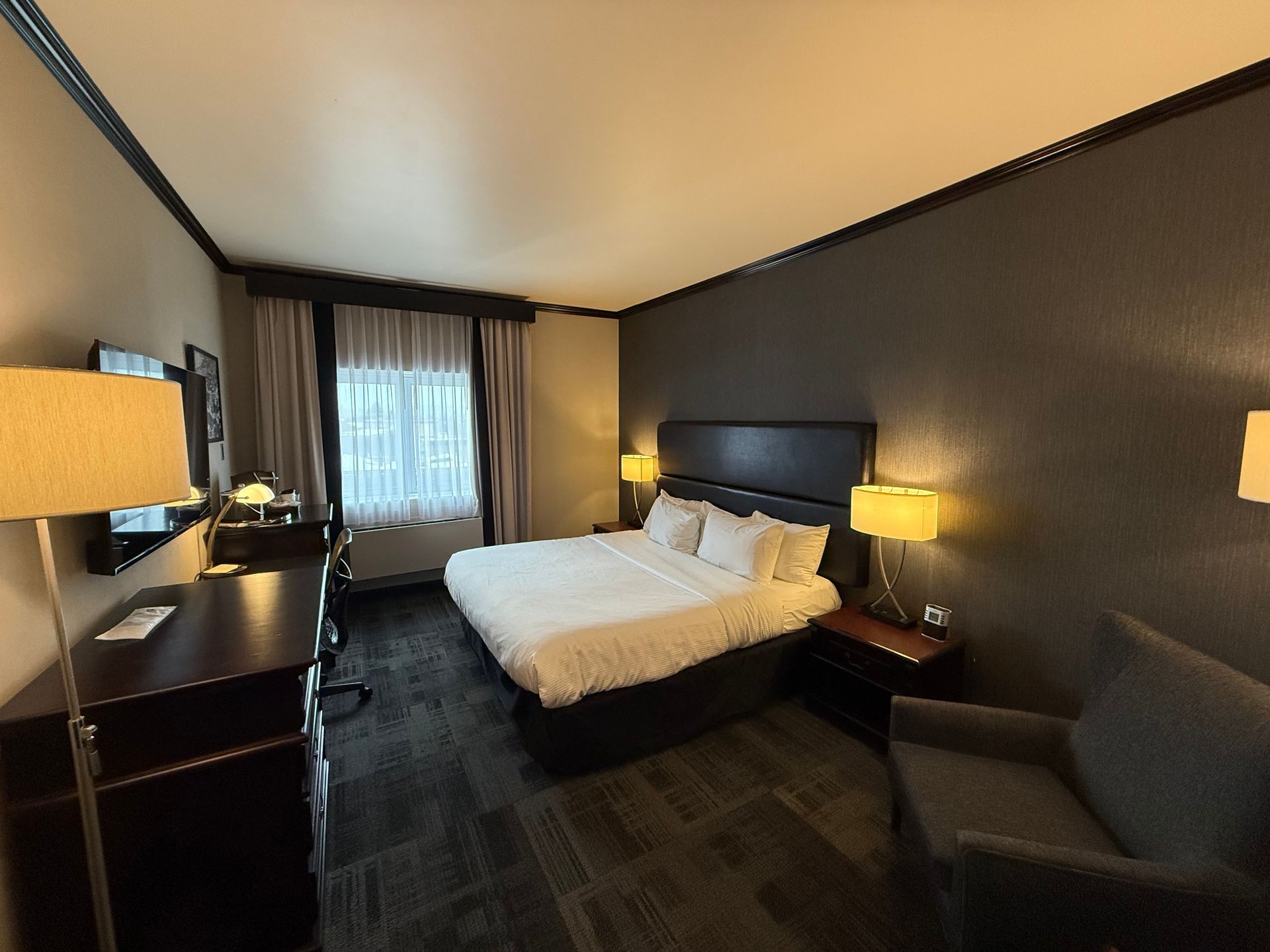 Chambre d'hôtel avec lit, bureau, chaise et table de chevet, éclairée par des lampes. 