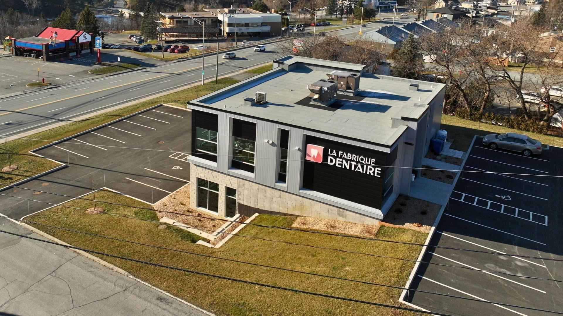 Vue extérieure d'une clinique dentaire moderne avec parking, espace vert et route visibles.