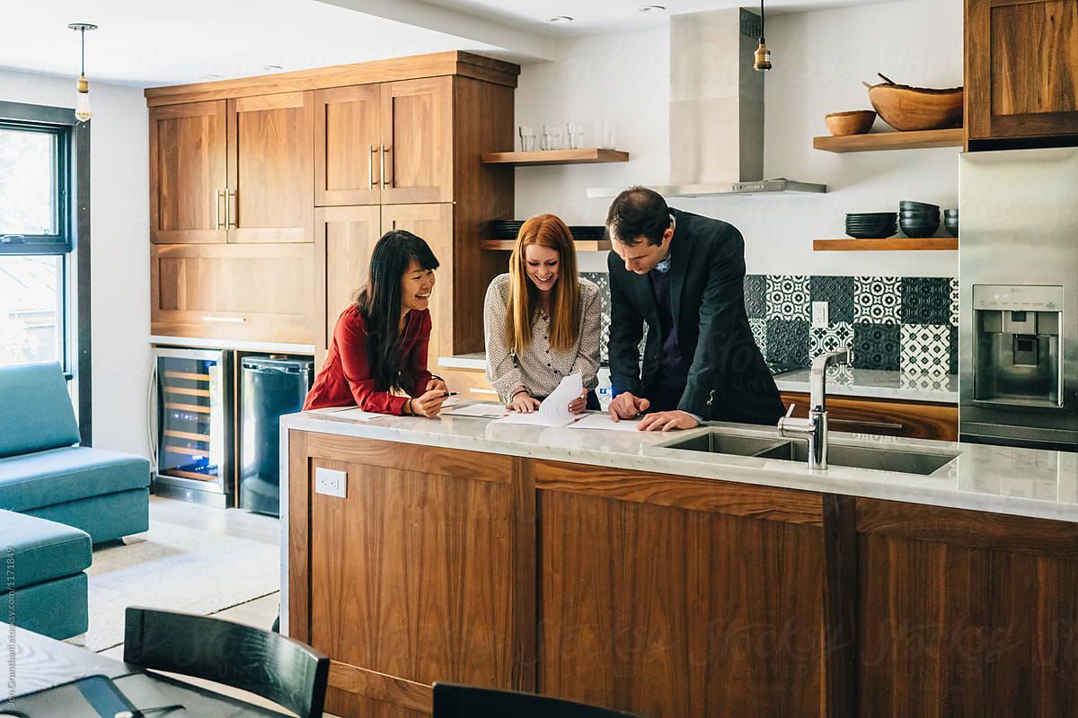 Three people reviewing documents at a kitchen island, discussing plans; wood cabinets.