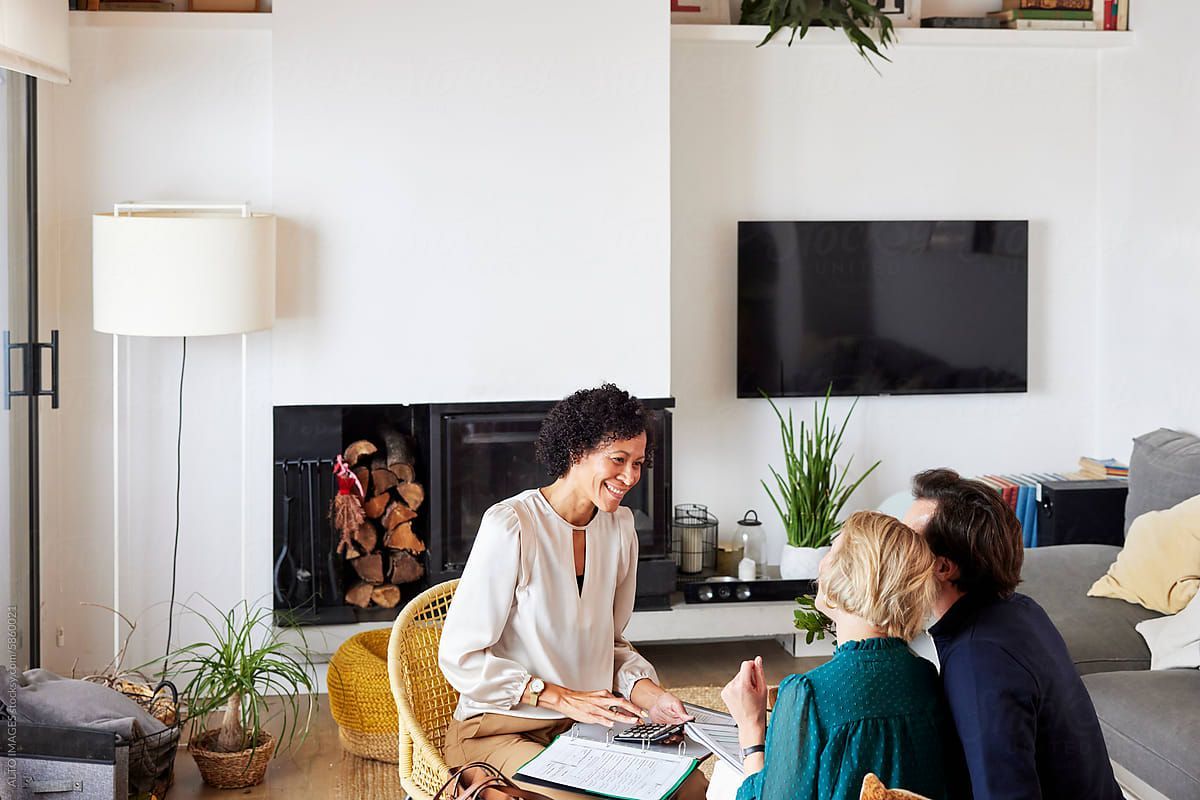 Woman consulting with couple in living room; smiling, discussing papers. Fireplace, plants, TV.