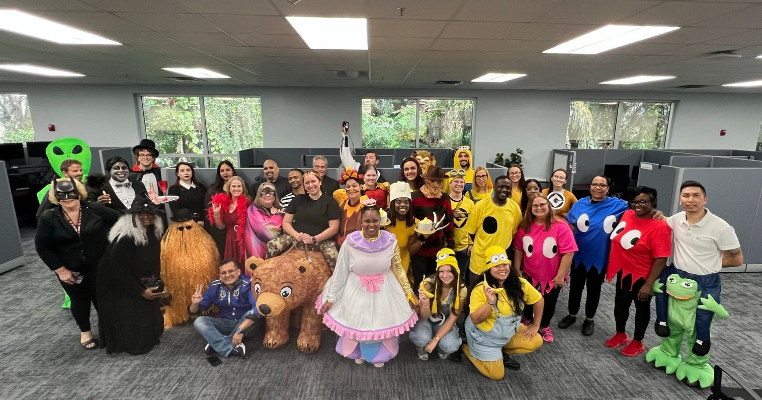 Office group in Halloween costumes, posing in front of cubicles. Many people smiling.