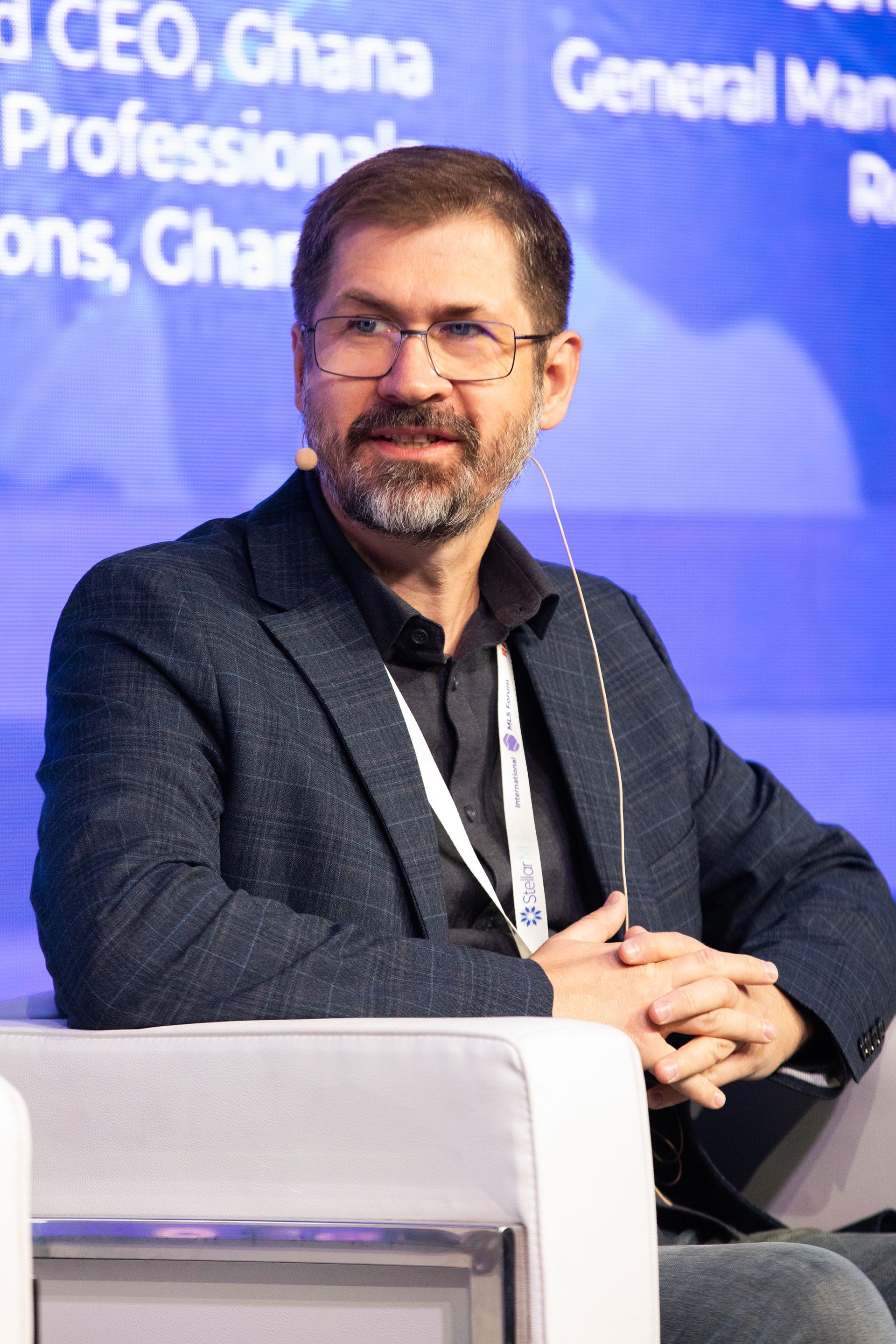 A man with a beard and glasses is sitting in a chair with his hands folded.