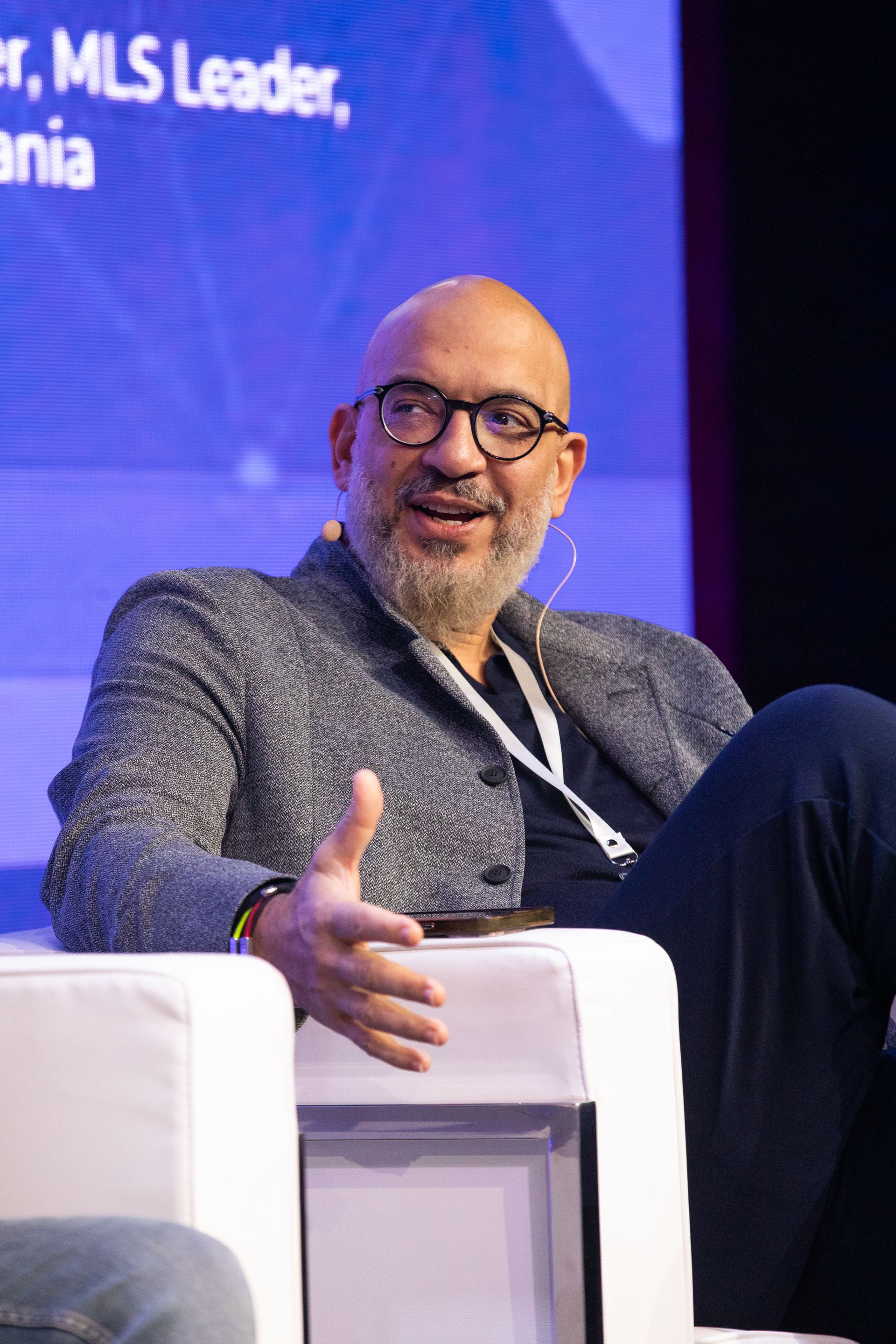 A bald man with glasses and a beard is sitting in a chair giving a thumbs up.