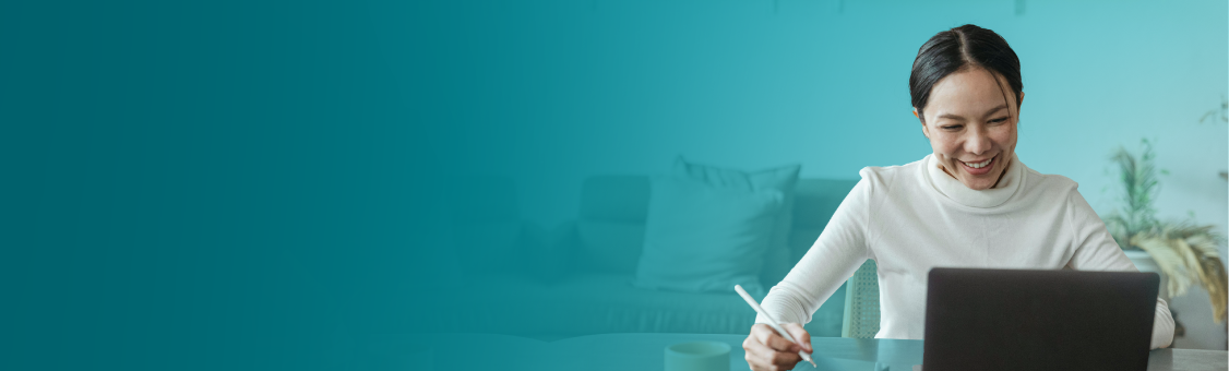 Woman smiling while using a laptop and writing with a pen at a table. Turquoise background.