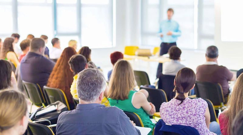 A group of people are sitting in a lecture hall watching a man give a presentation.