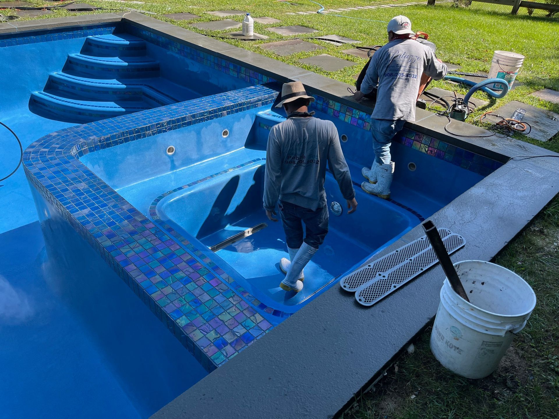Two workers wearing hats and grey clothing finish installing mosaic tiles on a blue swimming pool and attached spa.