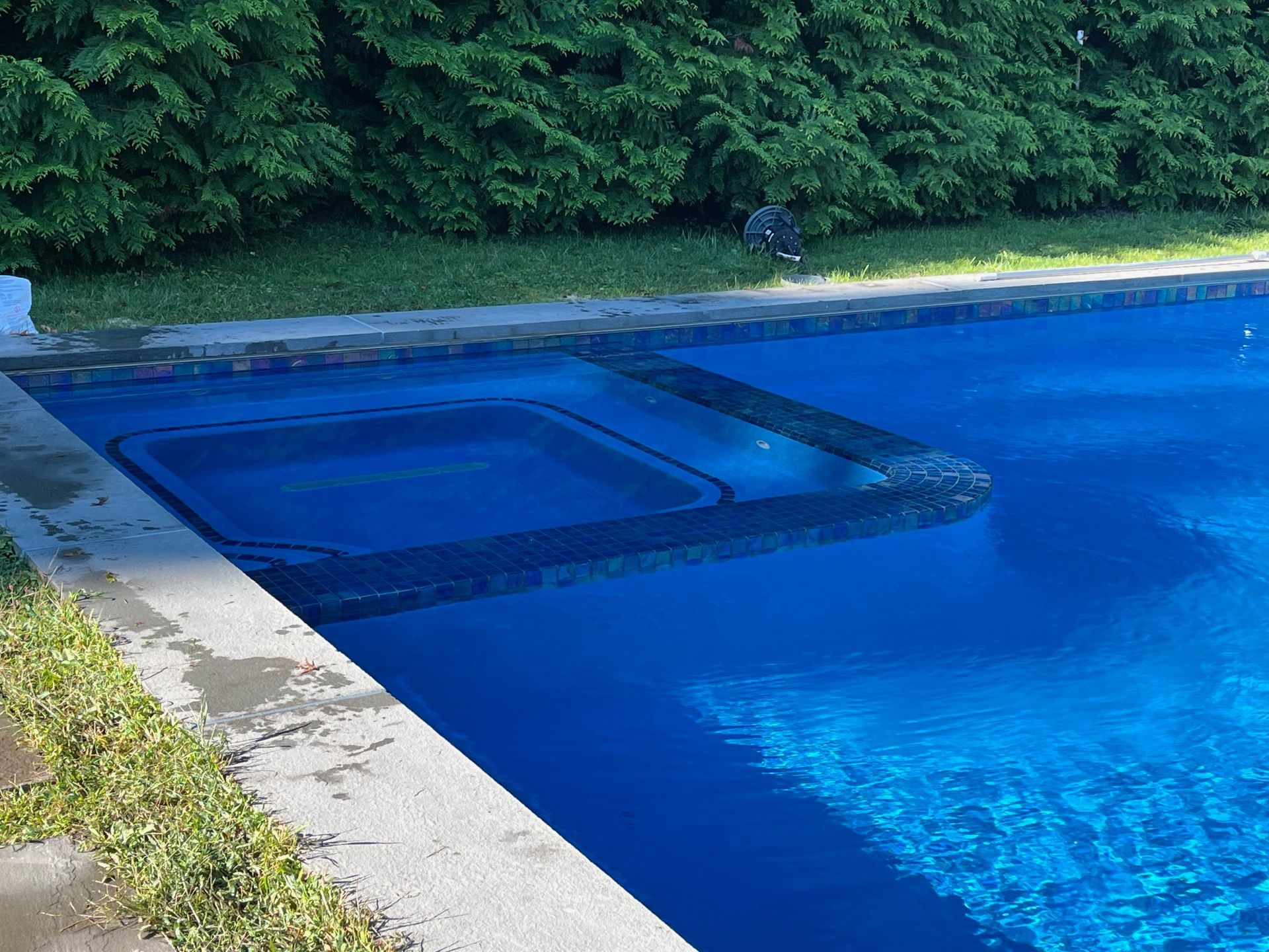 A rectangular, deep blue swimming pool features a built-in square spa surrounded by dark decorative tiling.