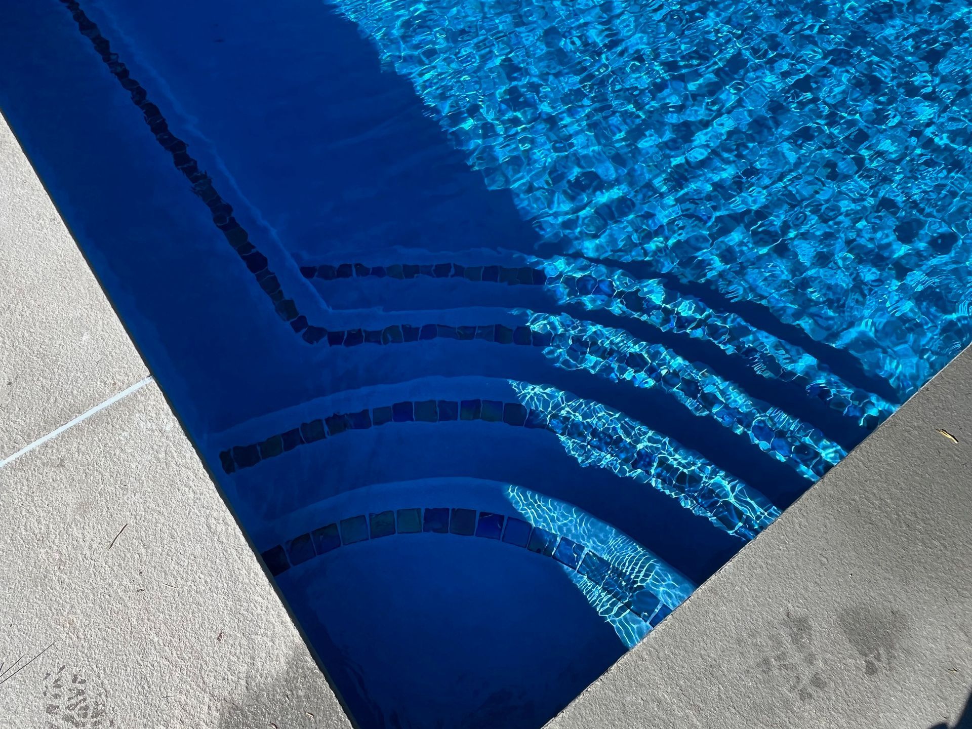 A top-down view of swimming pool stairs with dark tiled edges descending into vibrant blue water next to a concrete deck.